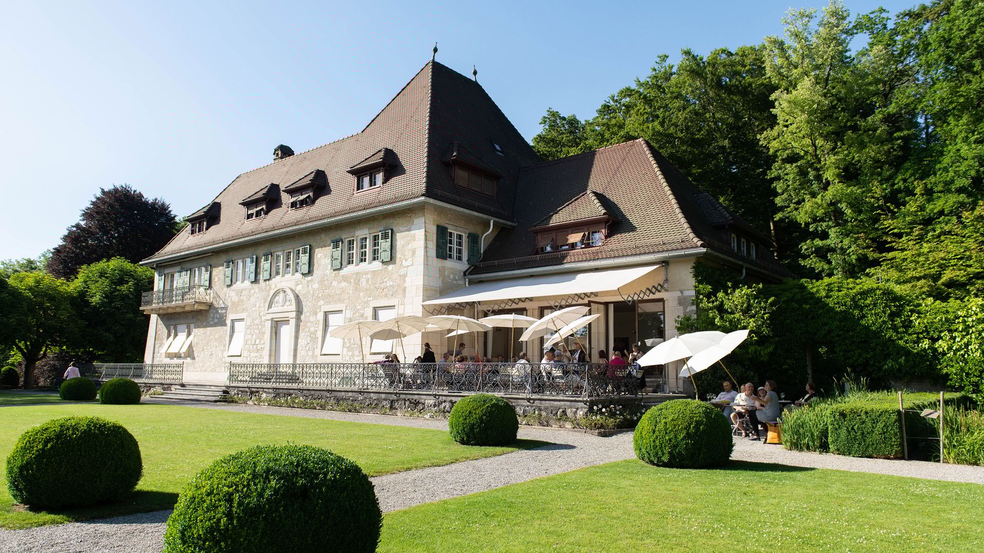 Grand bâtiment historique avec toit en pente et terrasse avec parasols devant pelouse soignée et buissons sphériques.