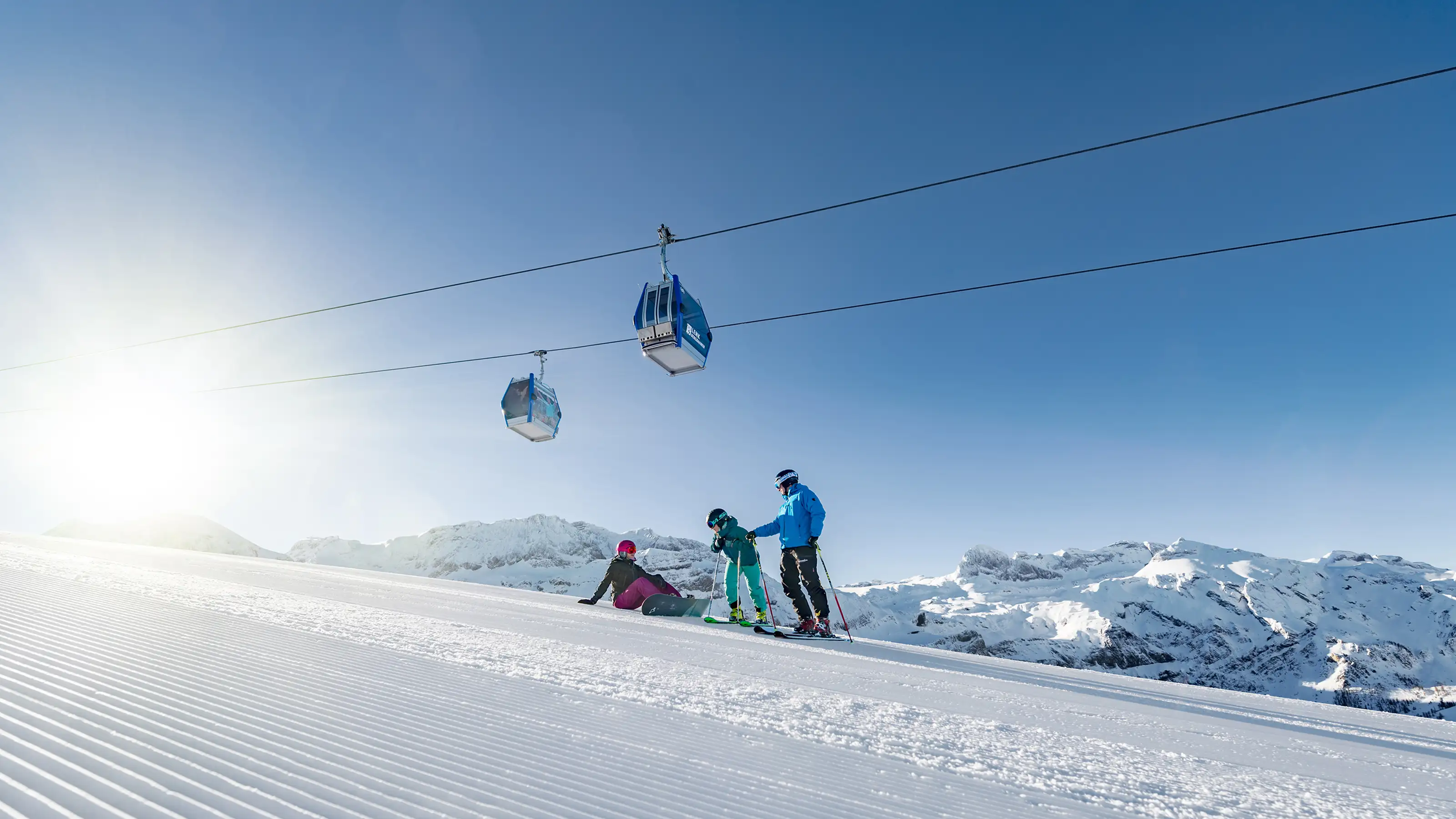 Skieurs en vêtements colorés sur piste damée dans la station Adelboden-Lenk, deux cabines de télécabine au-dessus, ciel ensoleillé.