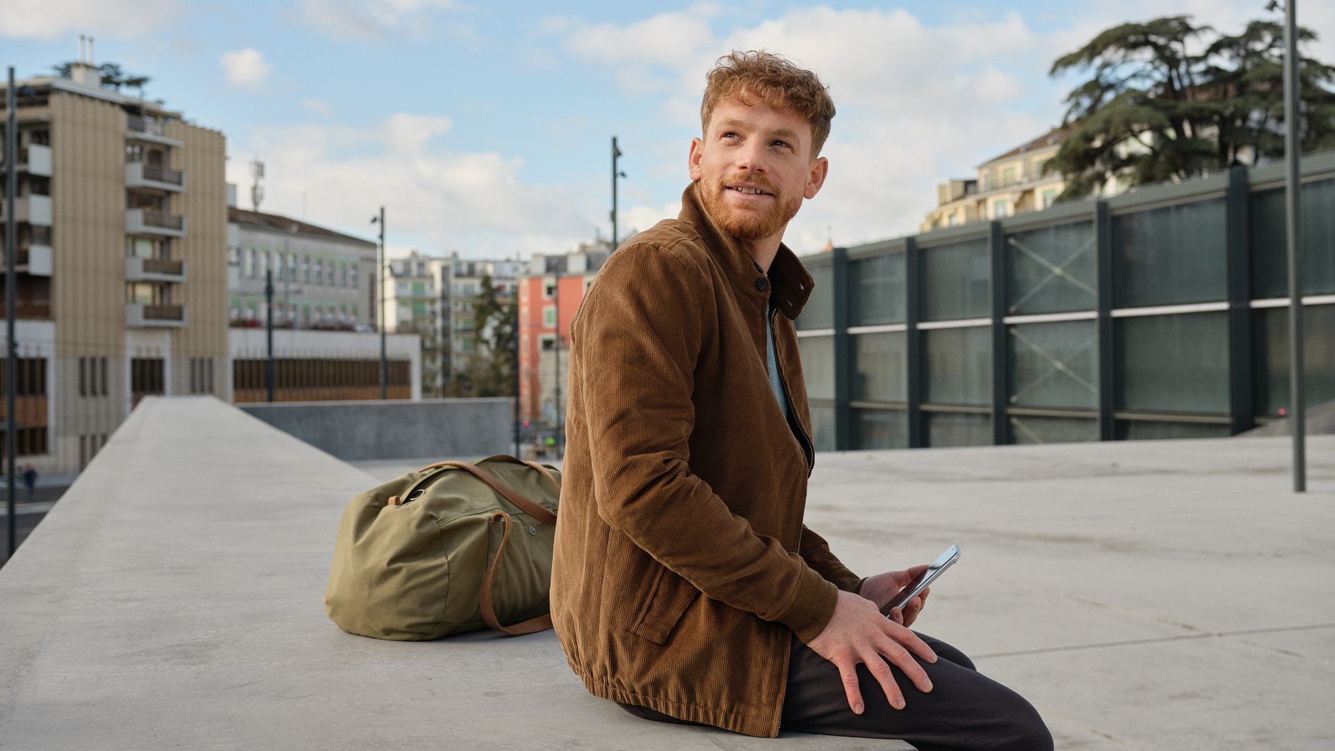 Un voyageur vêtu d'une veste marron est assis sur un mur devant la gare et tient un téléphone portable à la main.