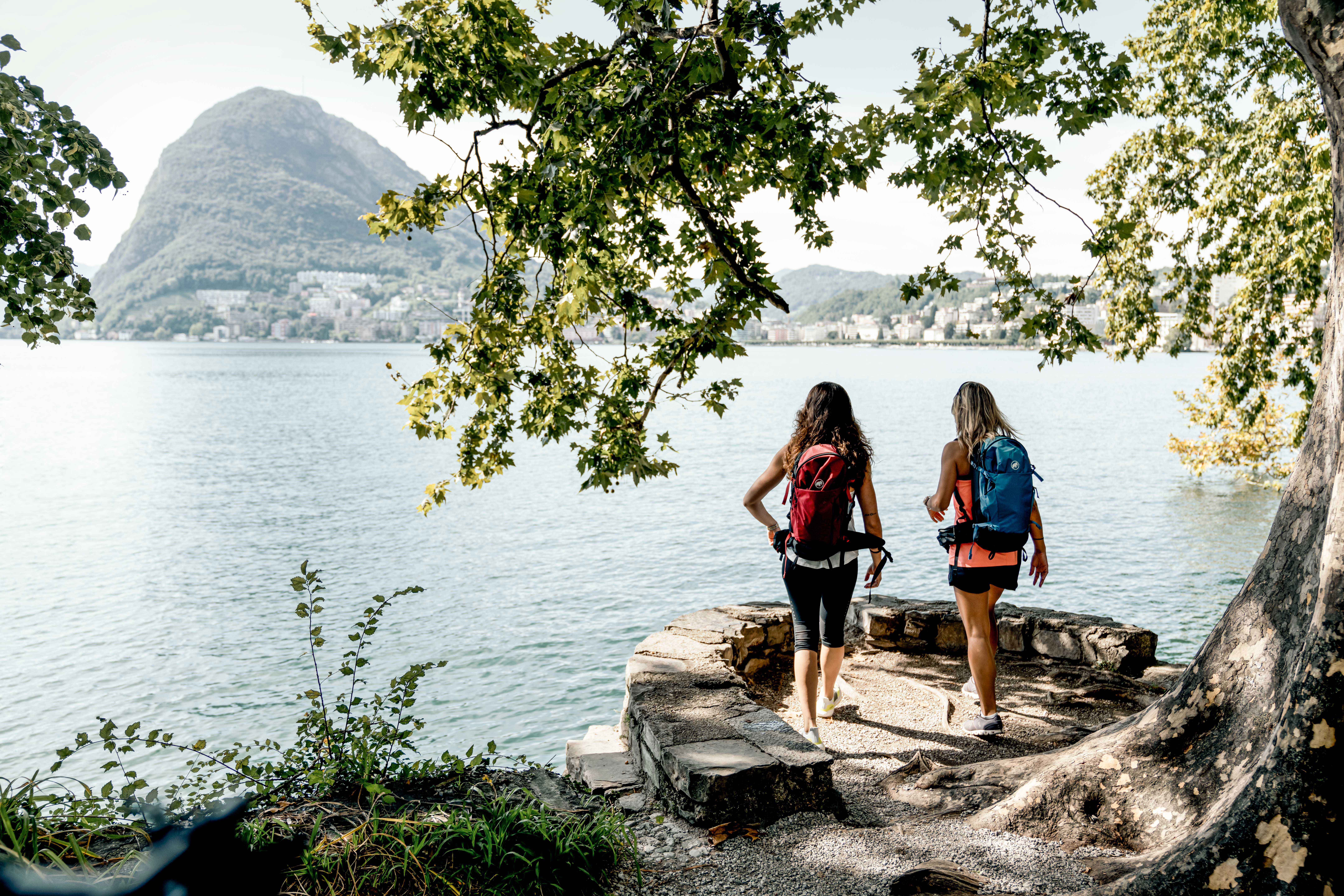 Zwei Frauen mit Rucksäcken gehen einen Weg entlang eines Sees.v