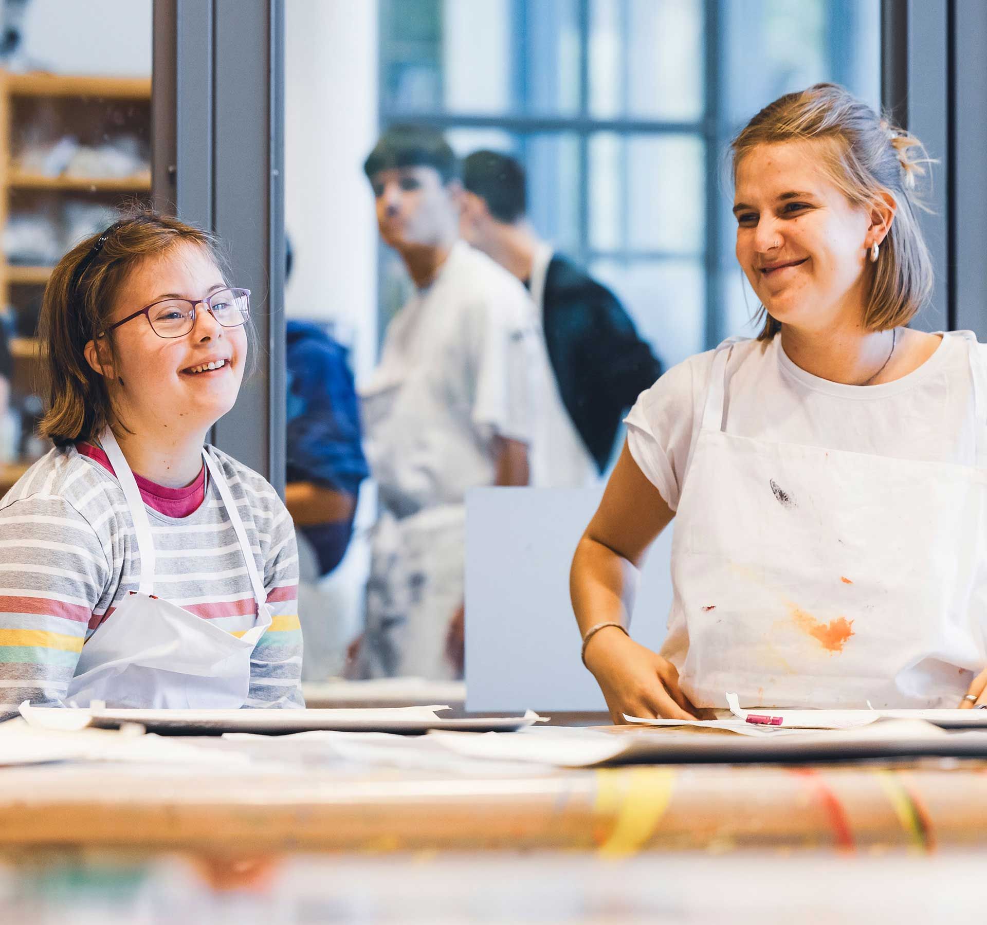 Two girls from art class smile at each other while surrounded by colourful art supplies.
