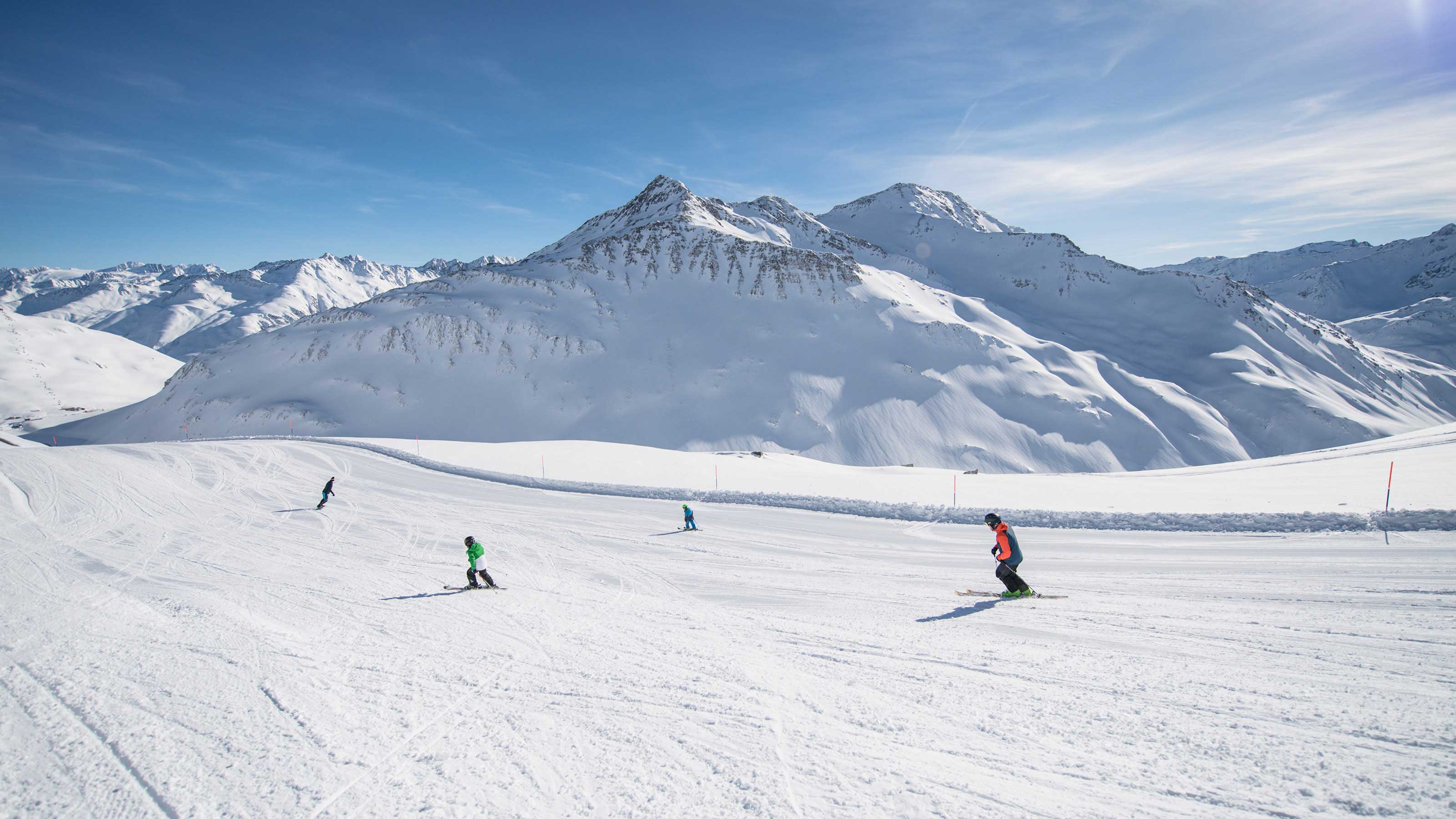 Sciatori su una pista ampia e battuta nell'area sciistica Andermatt-Sedrun-Disentis, montagne innevate sotto un cielo azzurro.