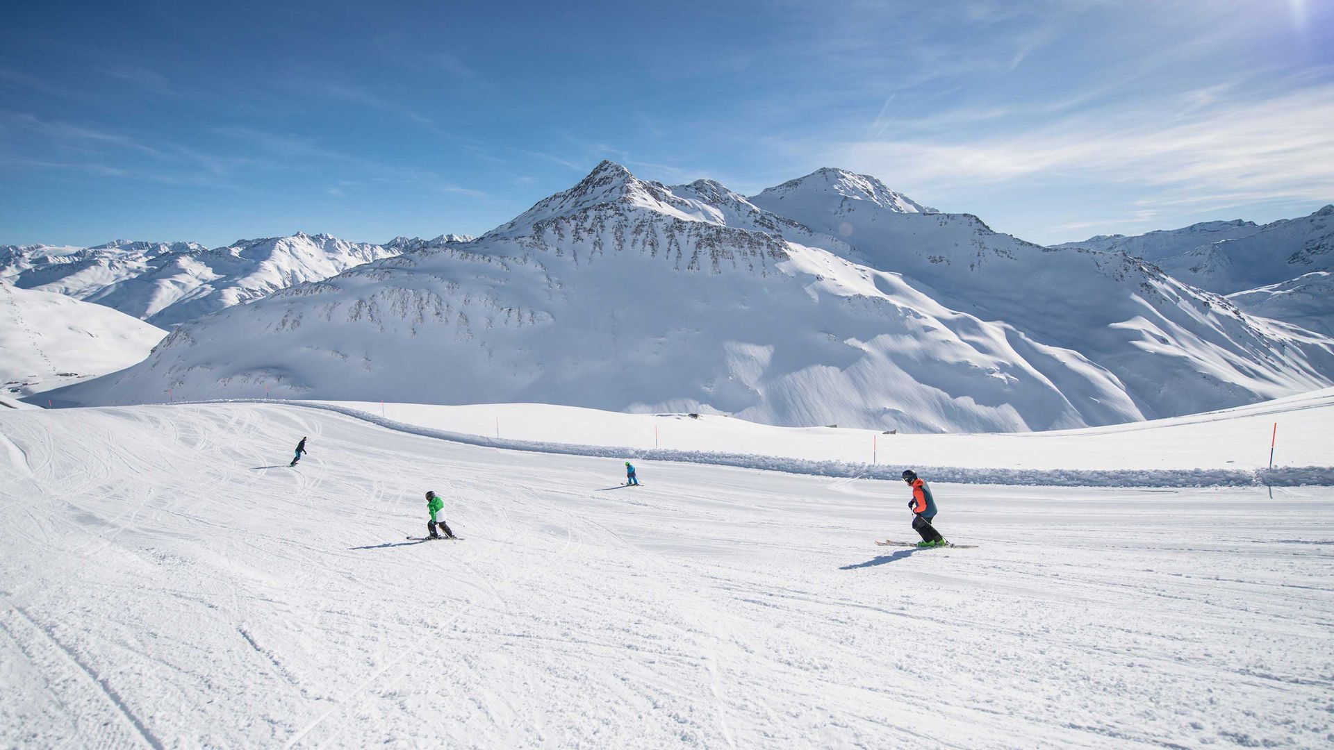 Skifahrer auf einer breiten, präparierten Piste im Skigebiet Andermatt-Sedrun-Disentis vor schneebedeckten Bergen unter blauem Himmel.