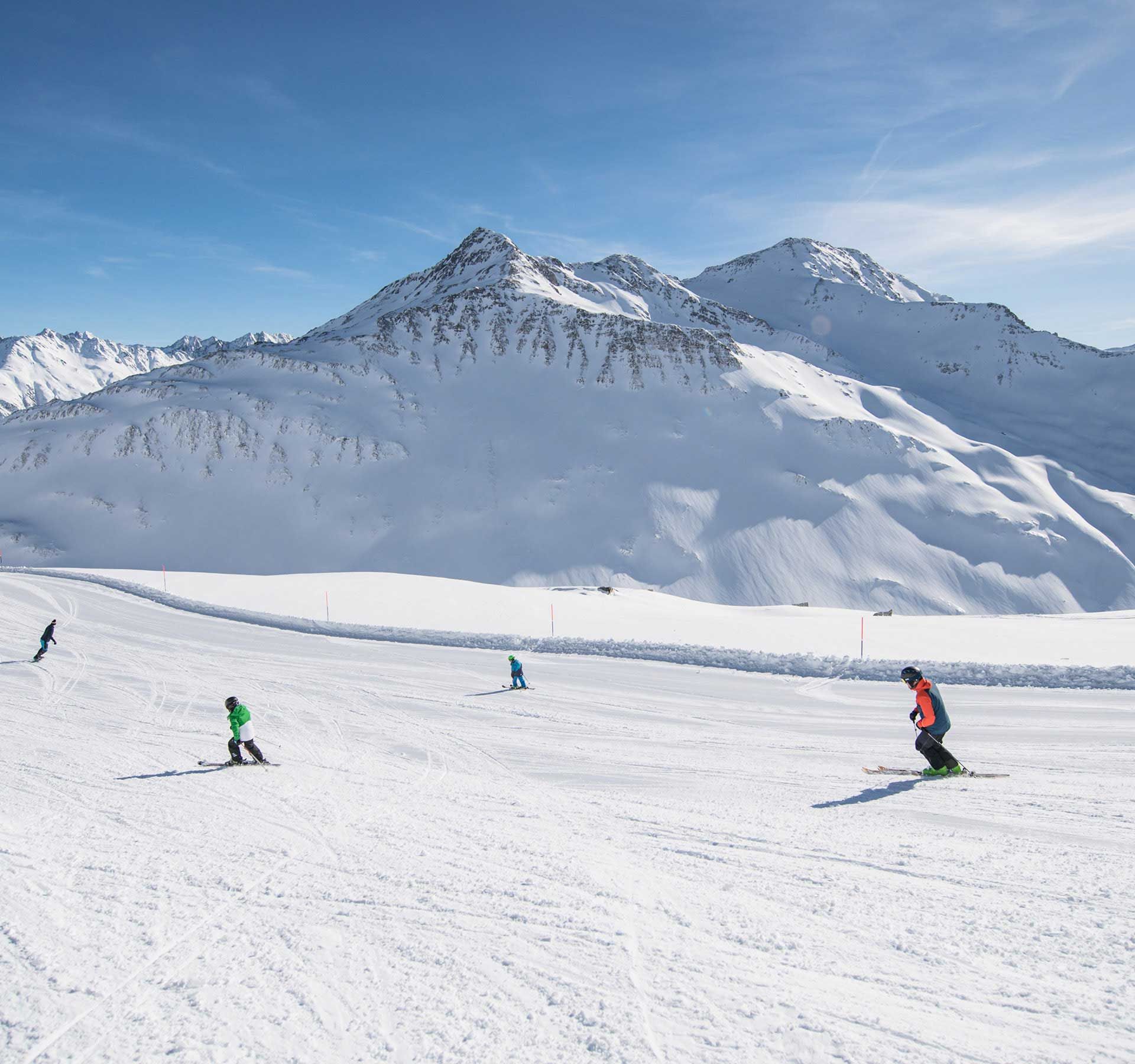 Skifahrer auf einer breiten, präparierten Piste im Skigebiet Andermatt-Sedrun-Disentis vor schneebedeckten Bergen unter blauem Himmel.