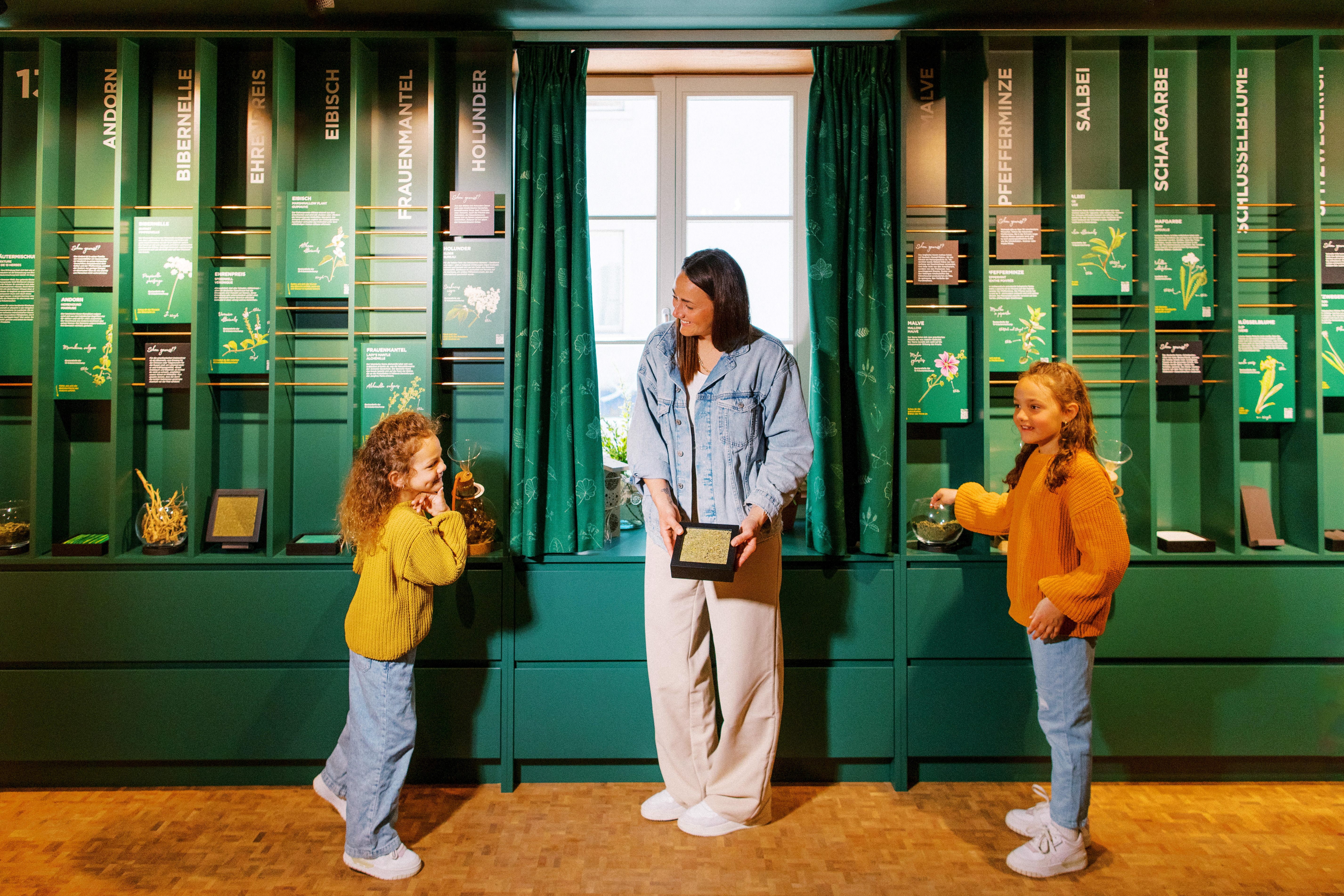 Una donna e due bambini ridono davanti a un muro con varie erbe.