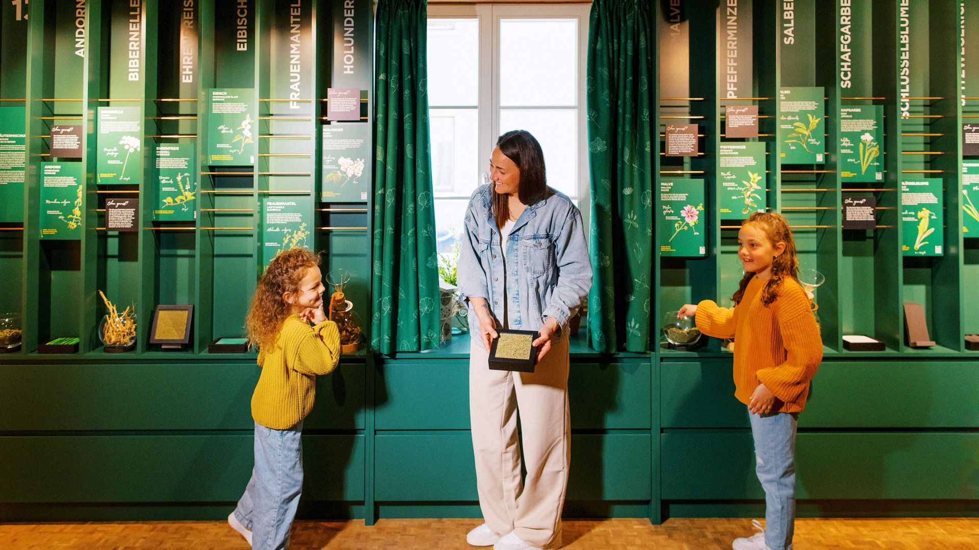 Eine Frau und zwei Kinder stehen lachend vor einer Wand mit verschiedenen Kräutern.