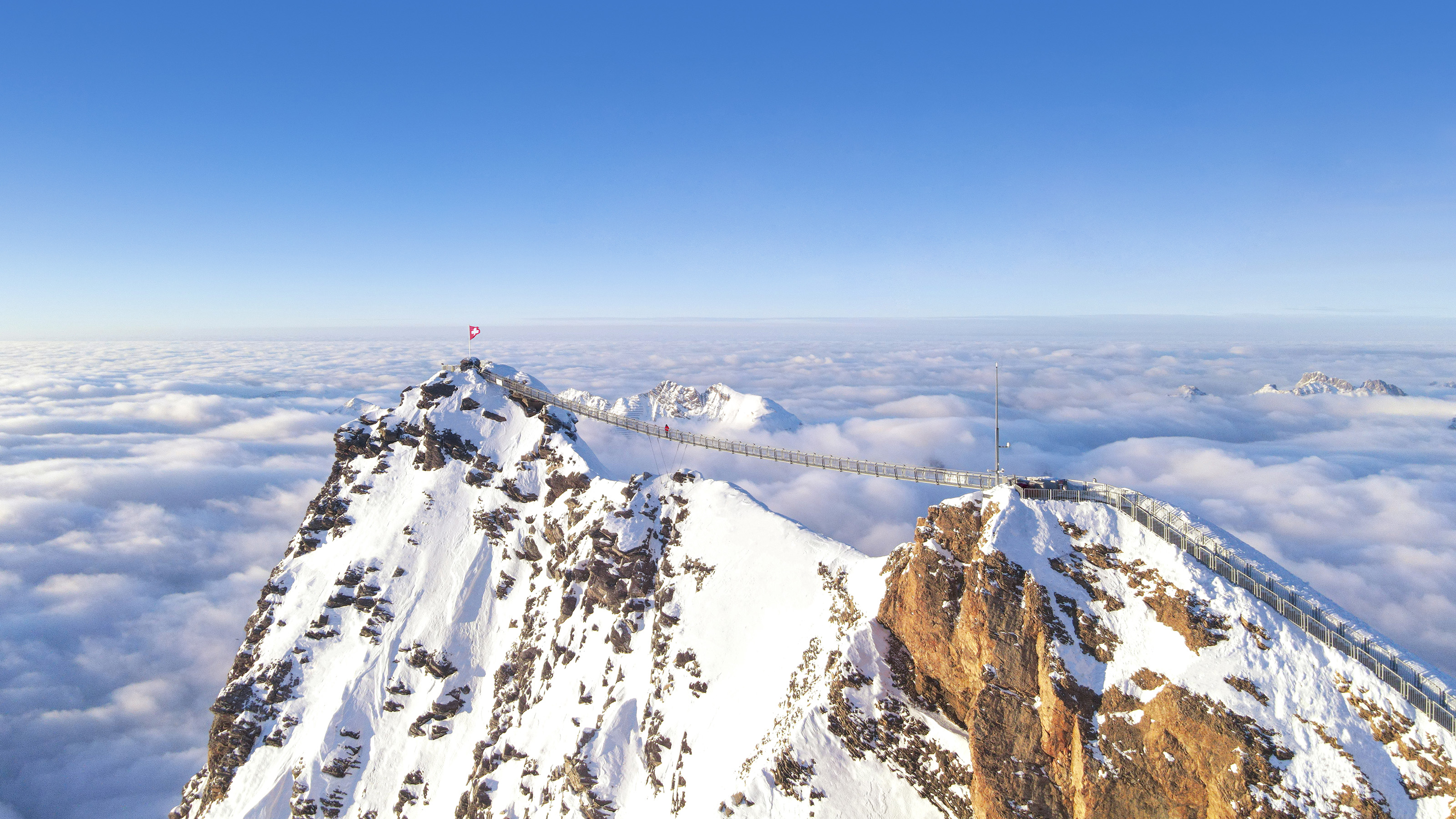 Luftaufnahme der Hängebrücke. Einzelne Gipfel ragen aus den Wolken hervor und werden von der Sonne in leuchtendes Licht gehült. 