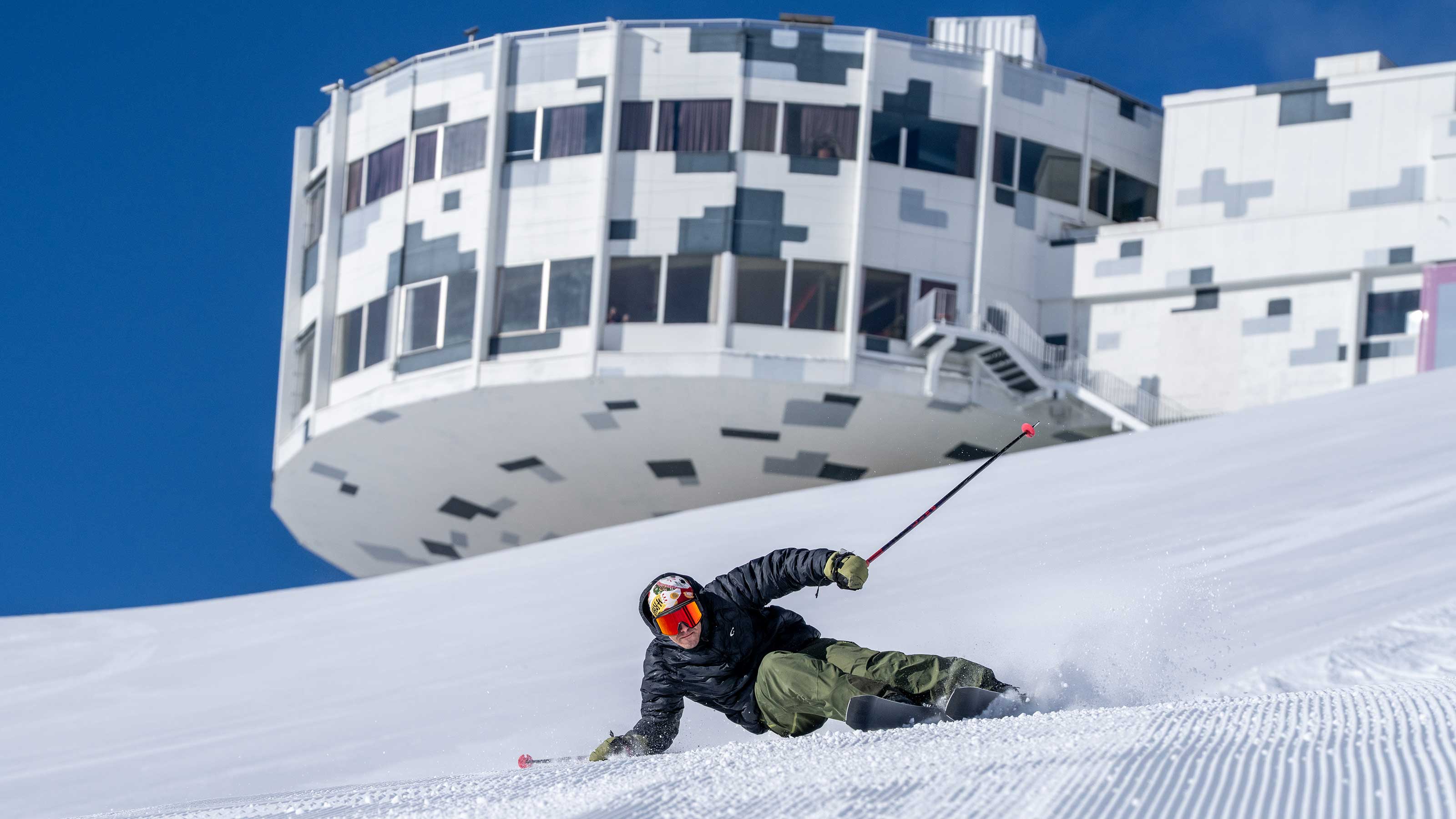 Skier wearing black jacket and green pants skiing on groomed slope in front of futuristic round building with white and black facade at Laax ski resort.