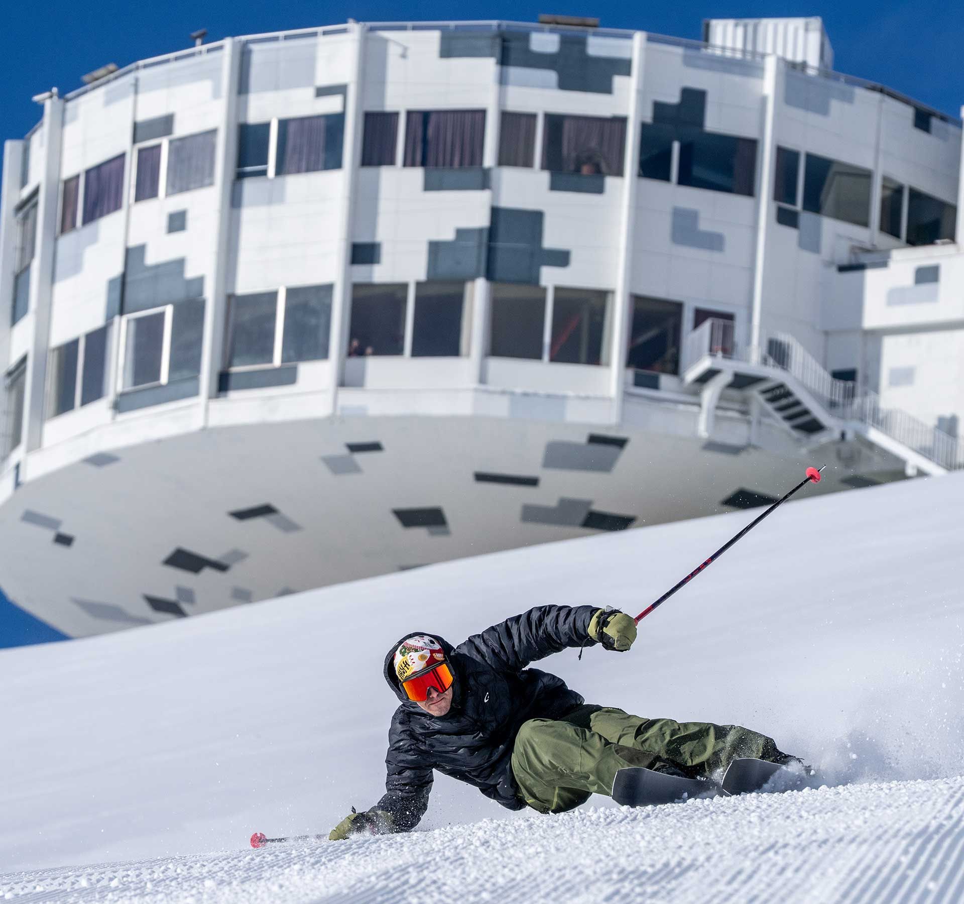 Skieur en veste noire et pantalon vert sur piste damée devant un bâtiment rond futuriste à façade blanche et noire dans la station de ski de Laax.
