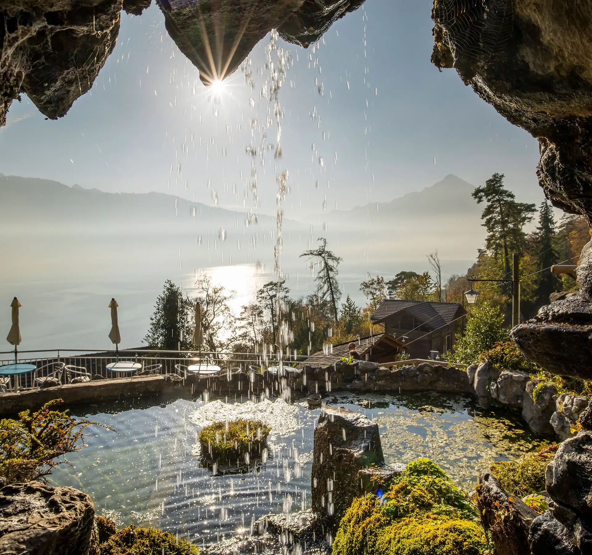 Vue depuis la grotte Saint-Beatus sur une cascade qui se jette dans un étang recouvert de mousse, avec un lac, des arbres et des montagnes en arrière-plan, sous le soleil.