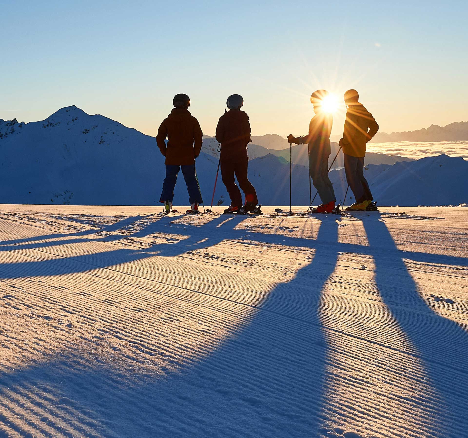 Vier Skifahrer stehen auf einer verschneiten Piste, blicken in die Ferne und geniessen den Sonnenaufgang.