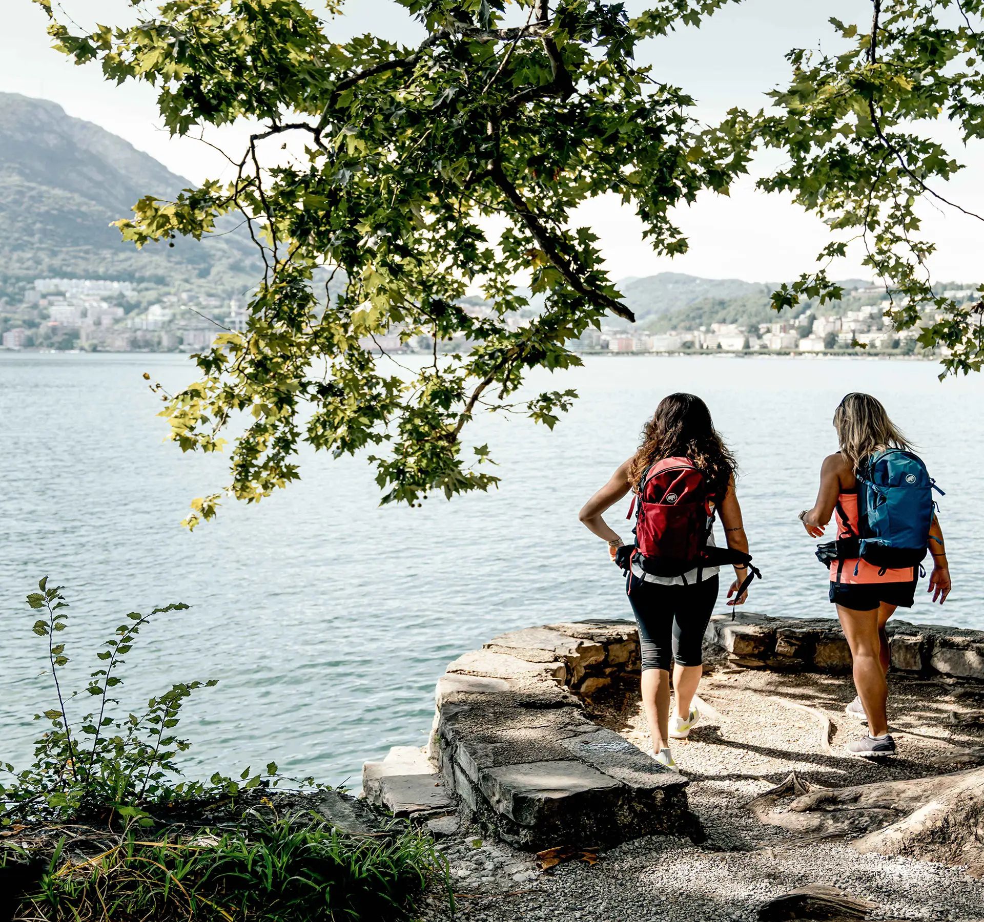 Zwei Frauen mit Rucksäcken gehen einen Weg entlang eines Sees.v