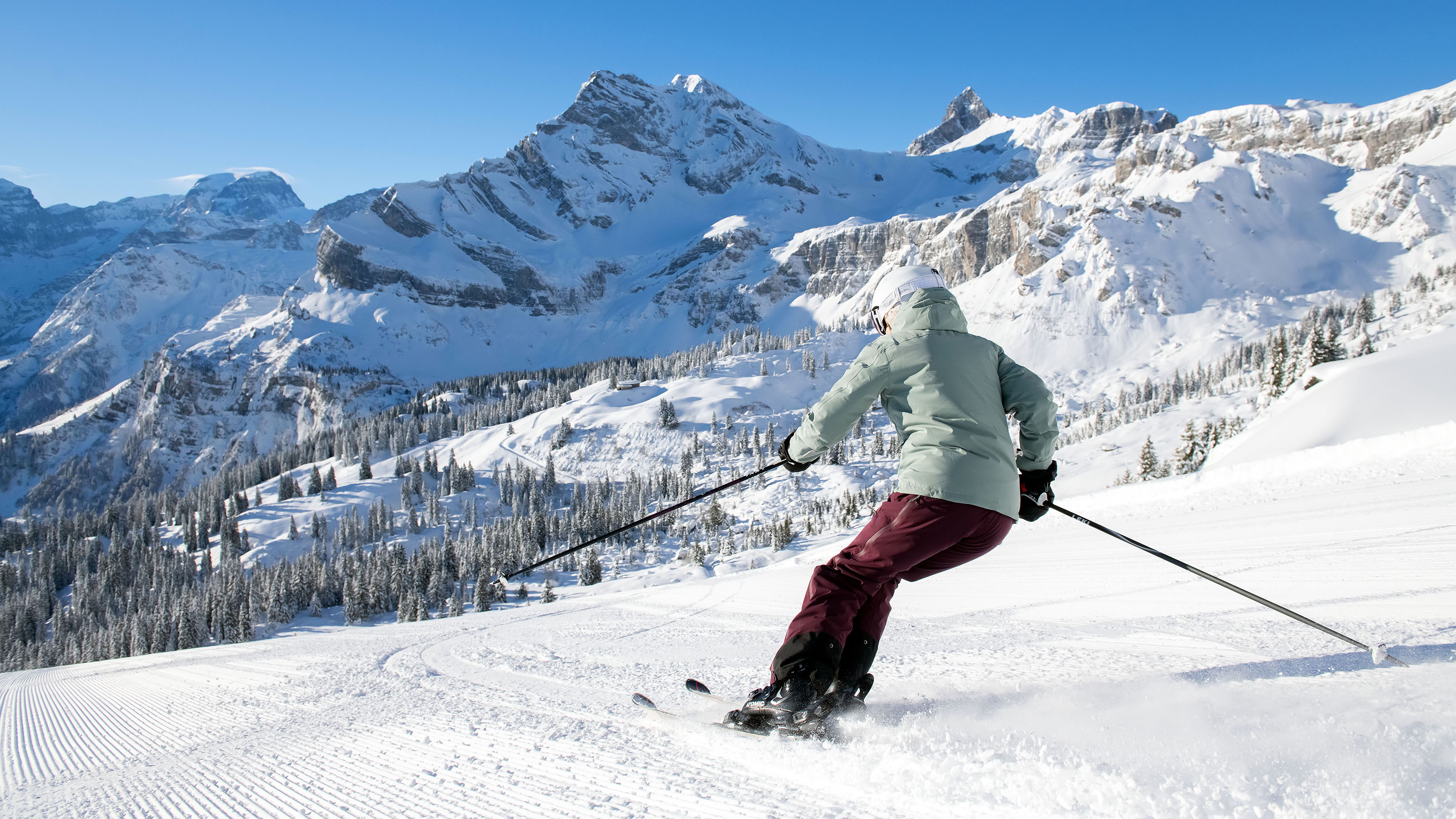 Skifahrerin in grüner Jacke fährt auf einer schneebedeckten Piste im Ski Gebiet Braunwald, umgeben von schneebedeckten Bergen und blauem Himmel.