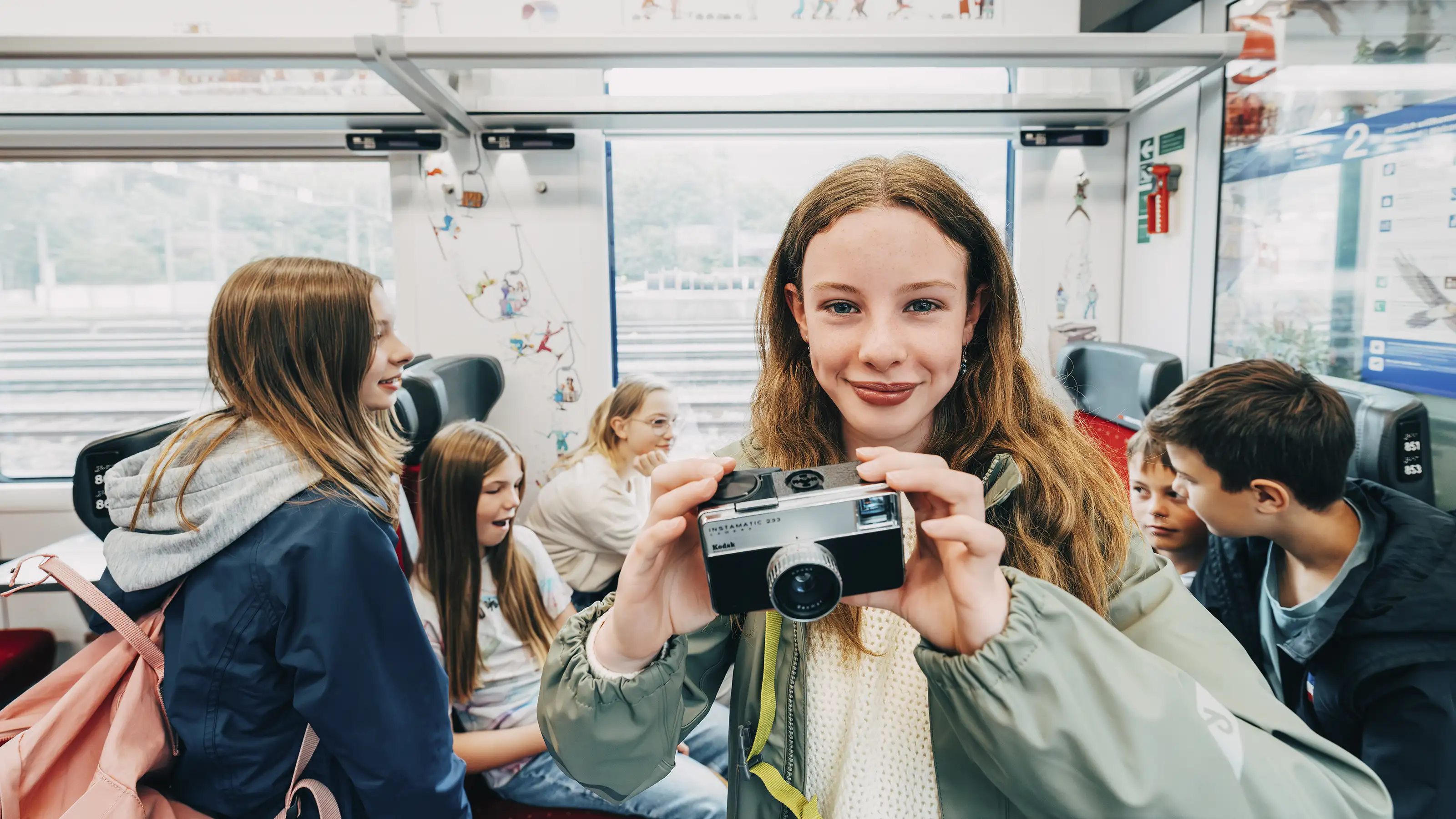 A girl stands in the train compartment and holds a camera into the picture.
