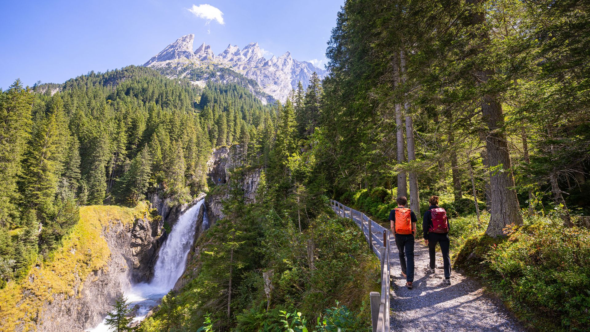Das Bild zeigt zwei Wanderer mit Rucksäcken auf einem Waldweg, der an der Rosenlaui Gletscherschlucht vorbeiführt, mit einer beeindruckenden Bergkulisse im Hintergrund.