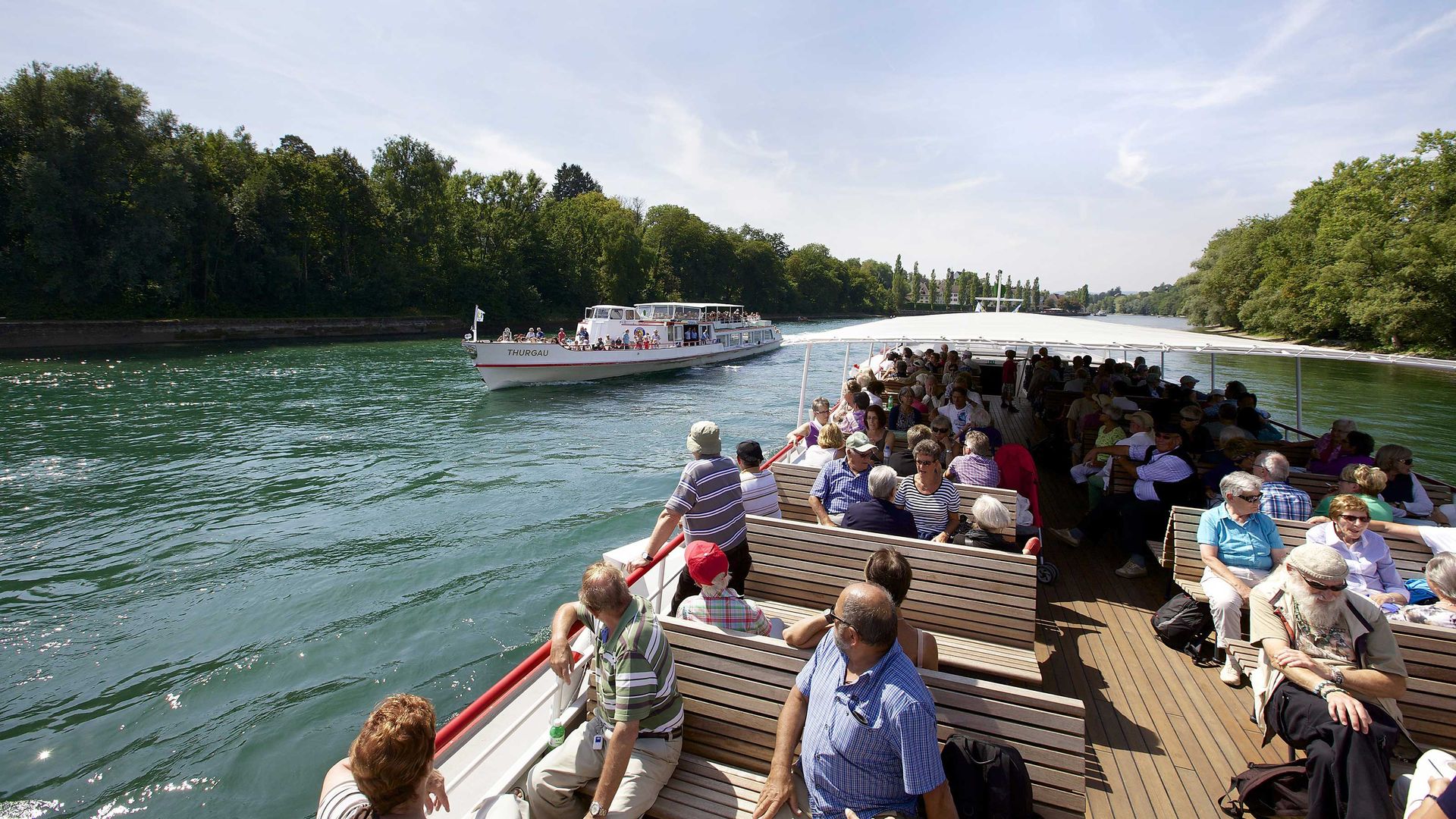A group of people sit relaxed at the back of a boat. In the background, a ship is travelling in the opposite direction.