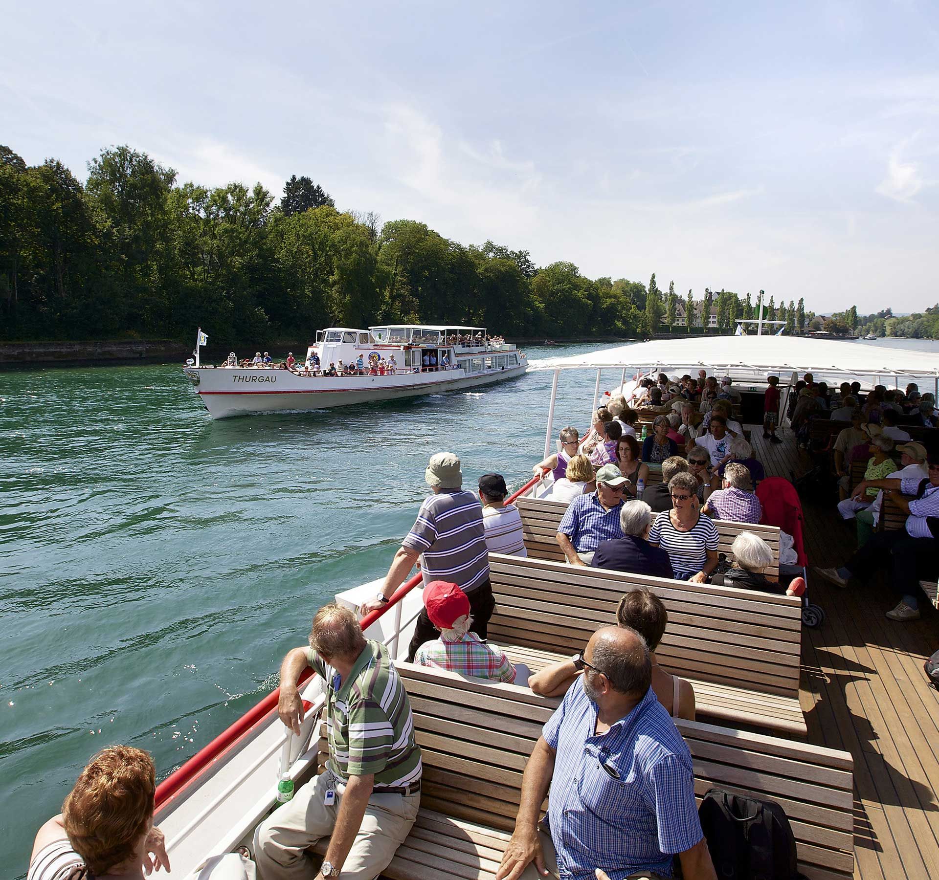 A group of people sit relaxed at the back of a boat. In the background, a ship is travelling in the opposite direction.