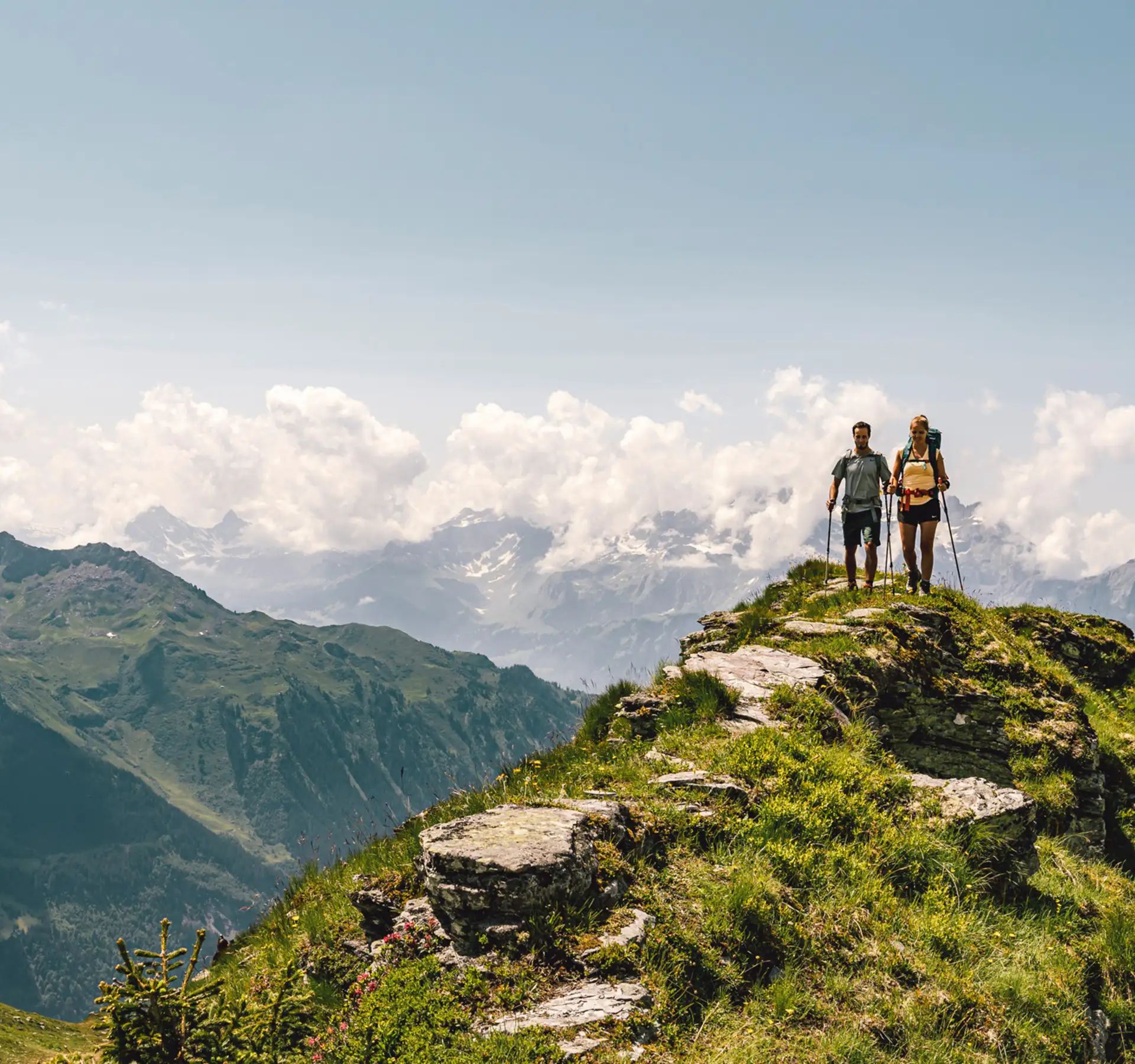 Man and woman hiking on a ridge in beautiful, sunny weather. In the background, numerous mountain peaks can be seen covered in clouds and partly covered in snow.