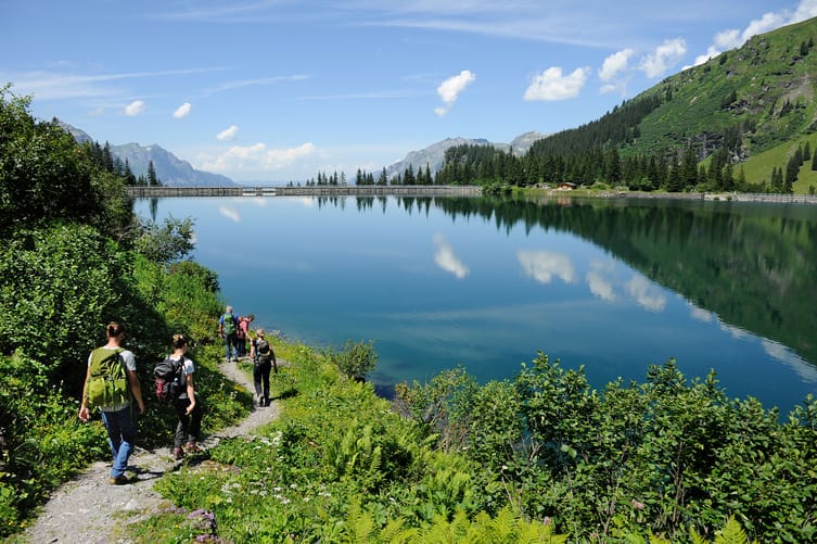 People are on a path that leads through the mountains next to a peaceful lake. 