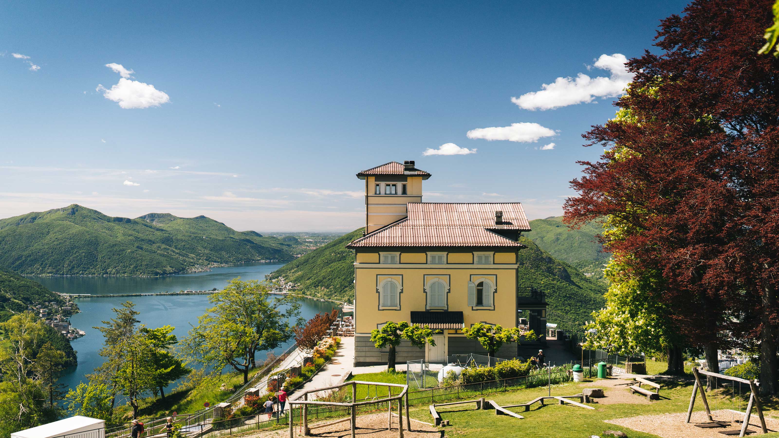 Un edificio giallo su una collina che domina il lago di Lugano, circondato da montagne verdi e alberi.