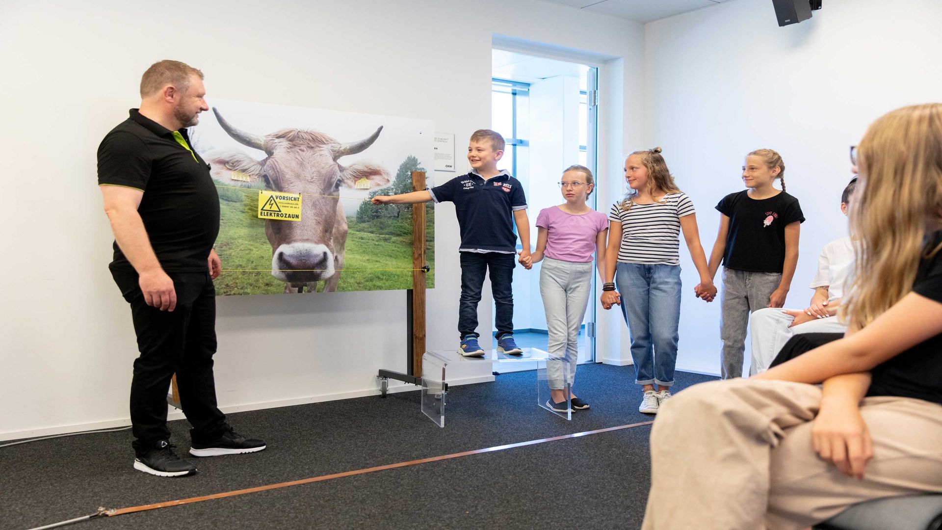 Eine Gruppe Schüler:innen stehen nebeneinander und halten sich an der Hand. Ein Schüler berührt in der CKW Energiewelt mit der Hand einen elektrischen Viehzaun um zu demonstrieren wie der Strom durch alle Schüler:innen dringt.
