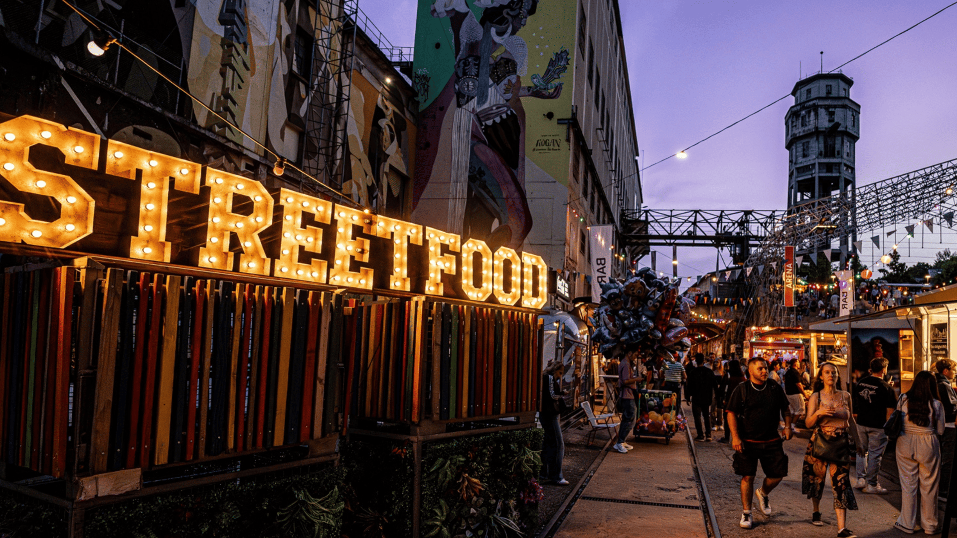 Des visiteurs se promènent dans un festival de street food avec des food trucks colorés, des guirlandes lumineuses et des stands de cuisine du monde entier.