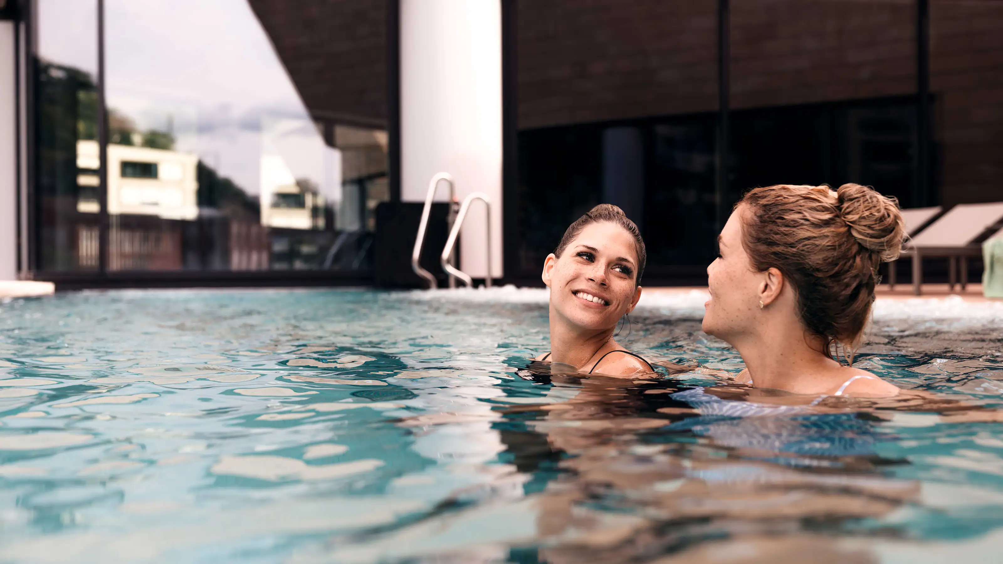 Two women enjoy a relaxing moment in a pool, surrounded by clear water and a sunny atmosphere.