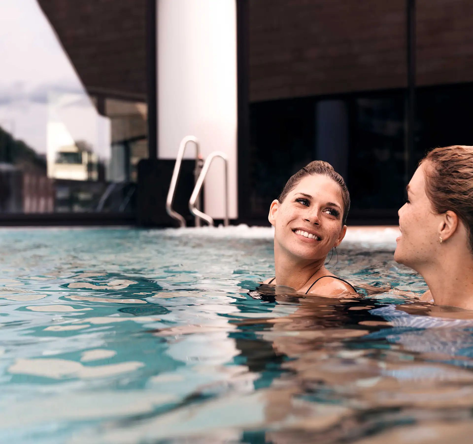 Zwei Frauen geniessen einen entspannten Moment in einem Hotelpool, umgeben von klarem Wasser und einer sonnigen Atmosphäre.