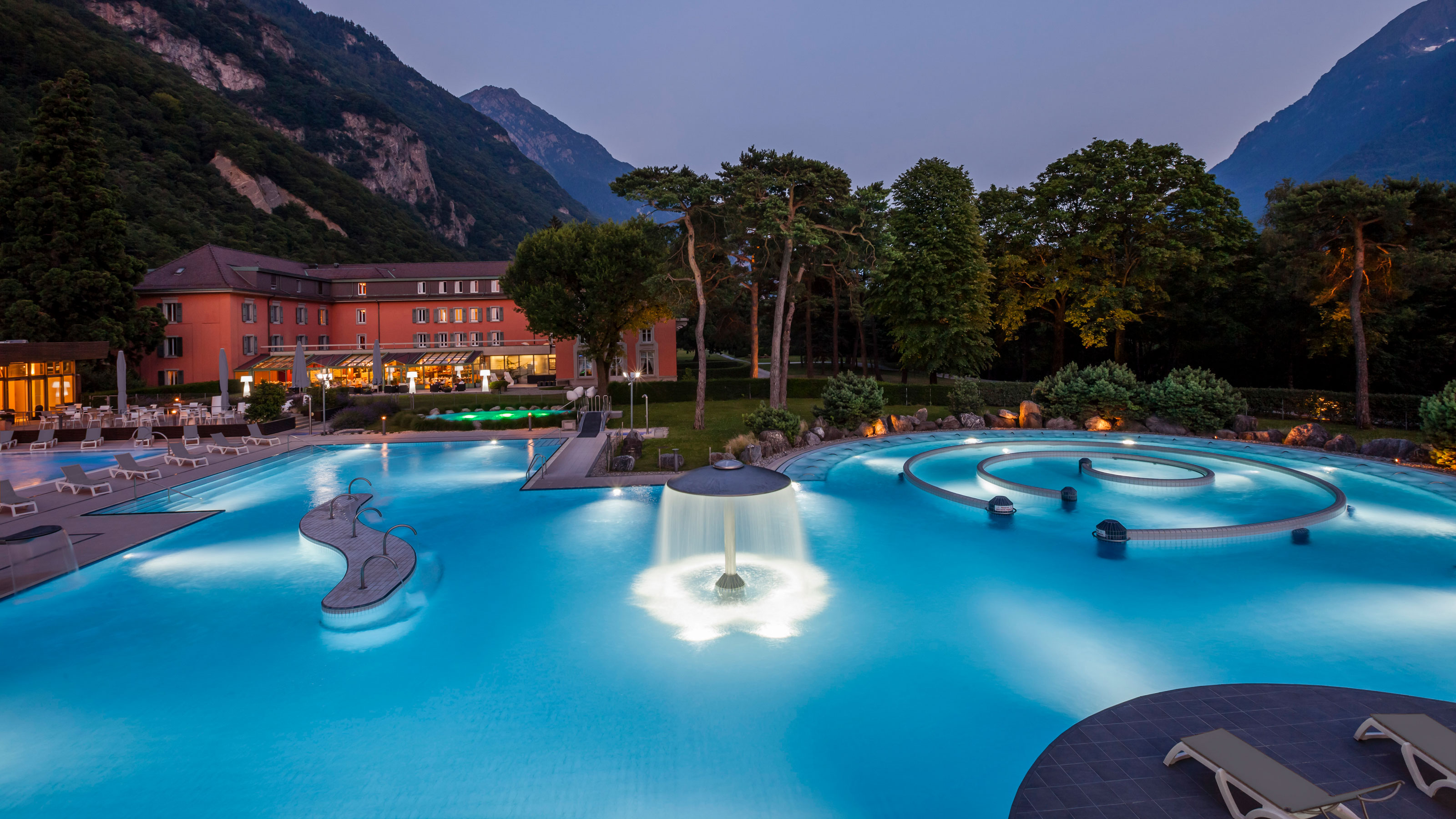 Une grande piscine extérieure avec le couché de soleil et des montagnes en fond.
