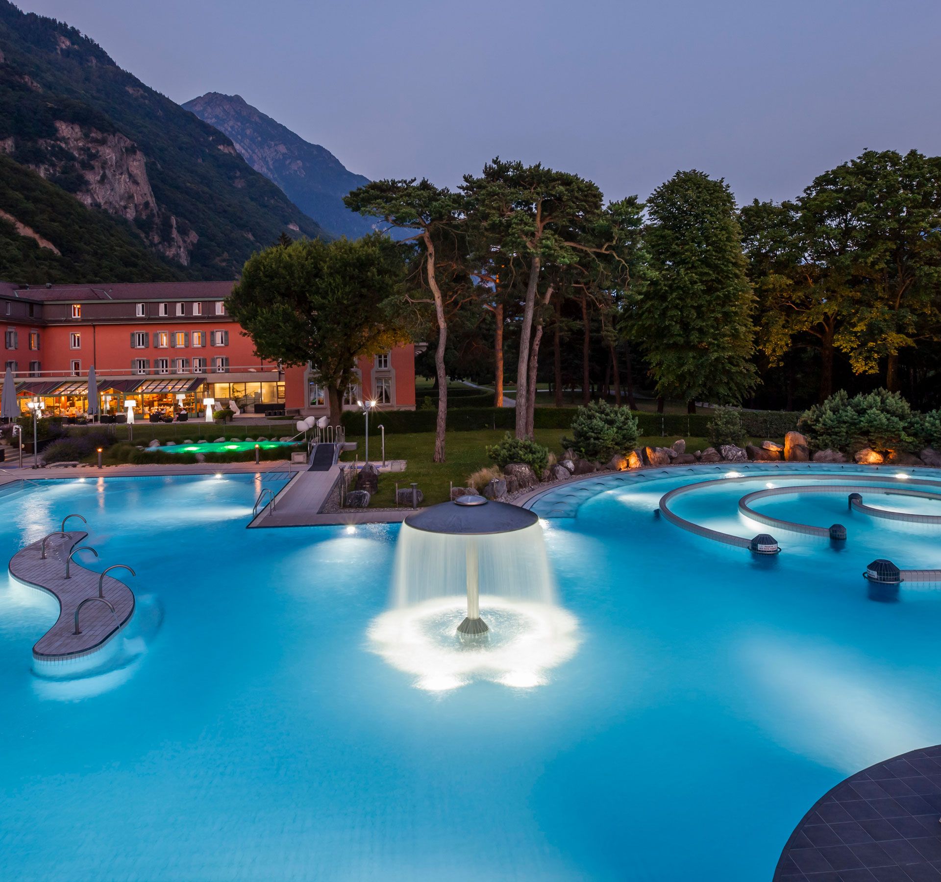 Une grande piscine extérieure avec le couché de soleil et des montagnes en fond.