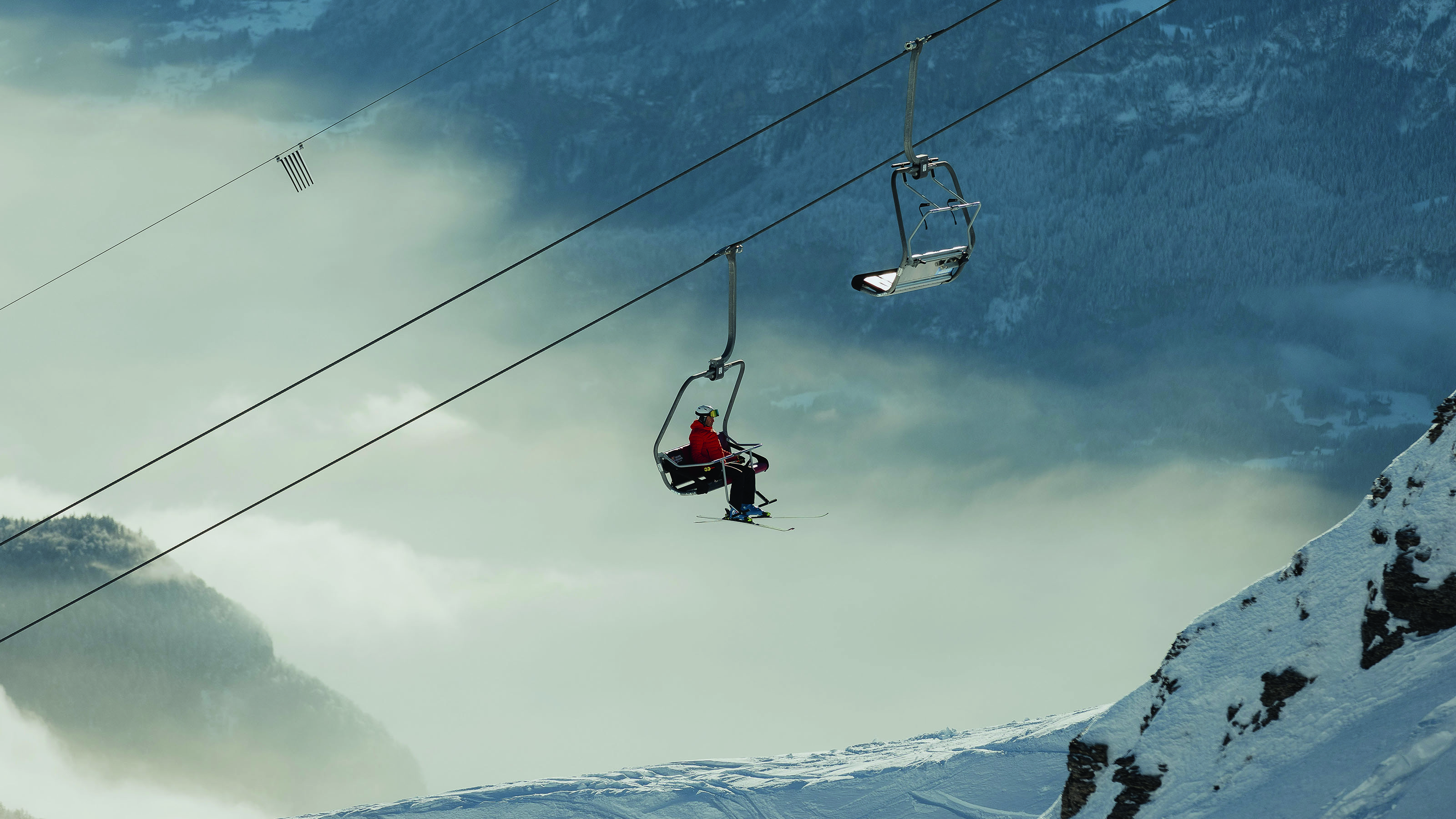 Chairlift in Sörenberg ski area with two people, one wearing a red jacket, above snowy mountain and clouds.