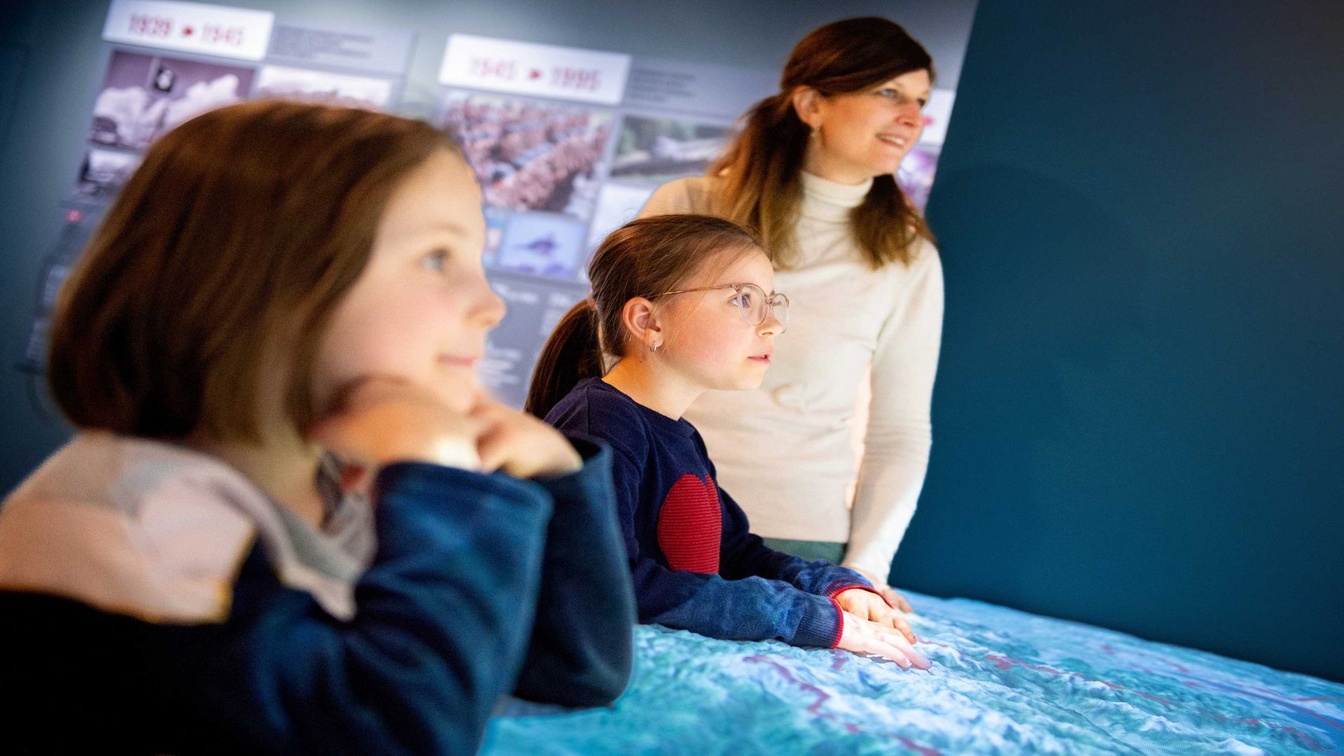 Eine Frau und zwei Kinder, die in einem Museum eine interaktive Ausstellung betrachten. Im Hintergrund sind Schautafeln mit historischen Bildern und Jahreszahlen zu erkennen.