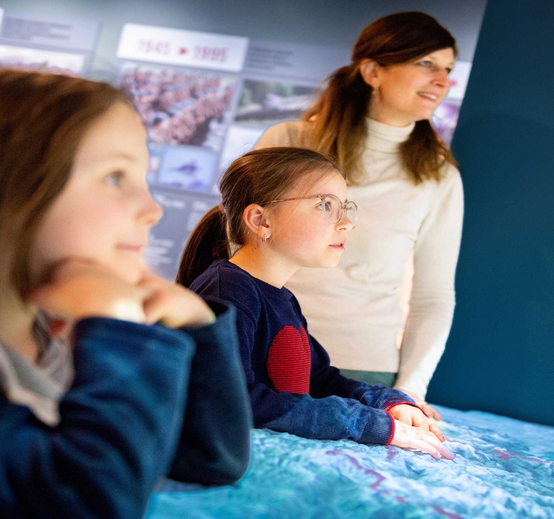A woman and two children looking at an interactive exhibition in a museum. Display boards with historical pictures and dates can be seen in the background.