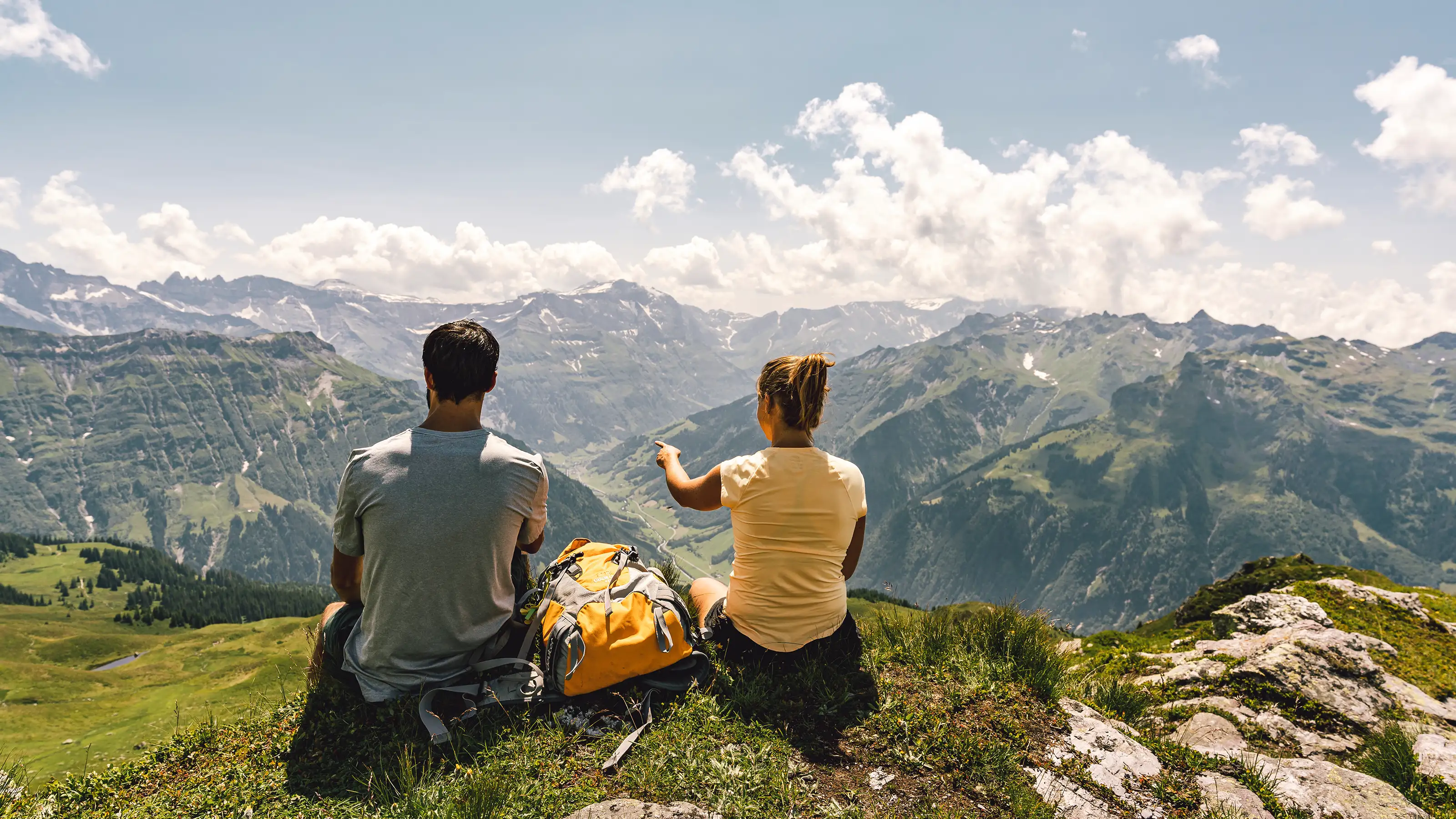 Due persone sono sedute a terra e si godono insieme una splendida vista sul mondo alpino.