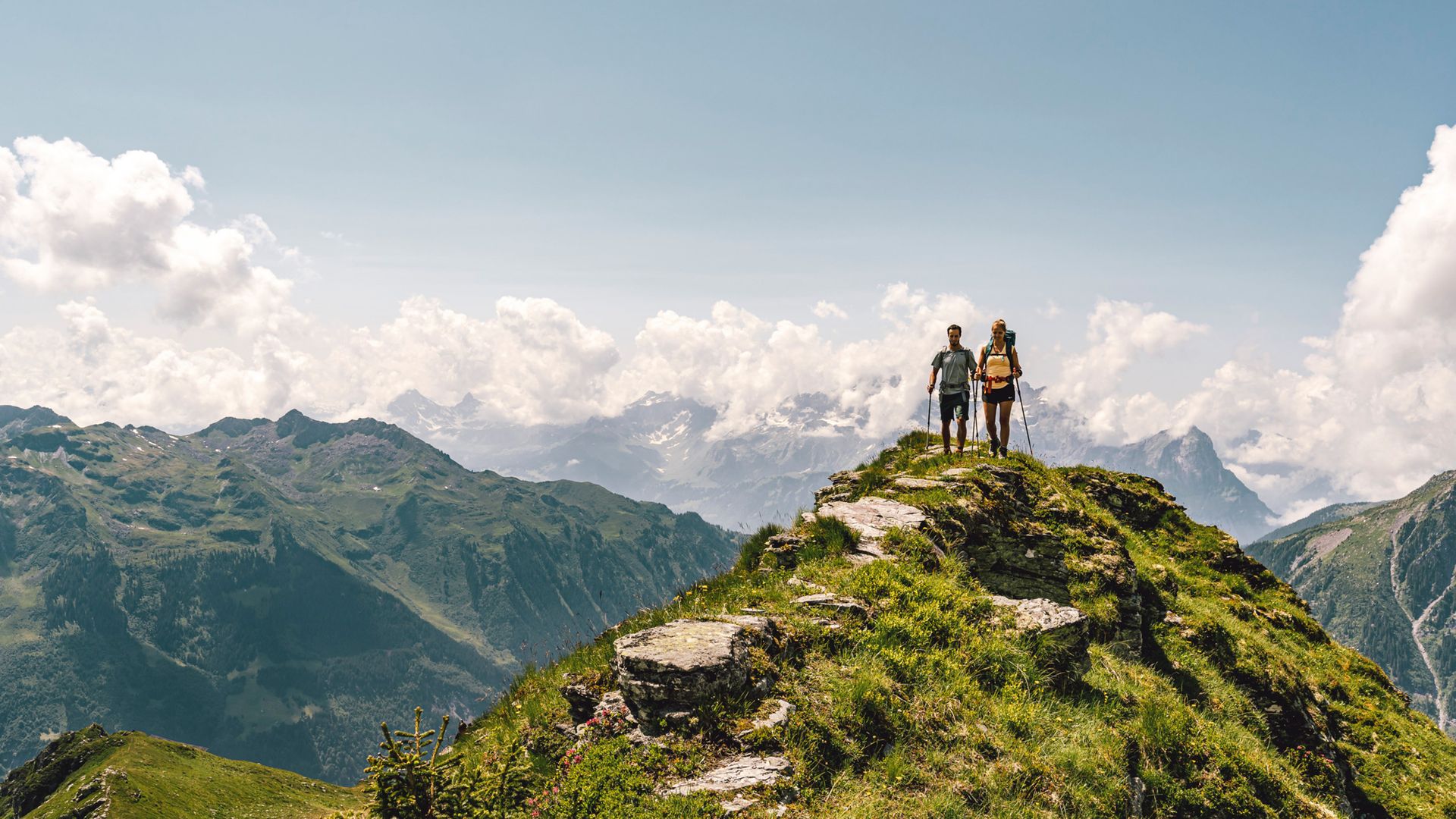 Man and woman hiking on a ridge in beautiful, sunny weather. In the background, numerous mountain peaks can be seen covered in clouds and partly covered in snow. 