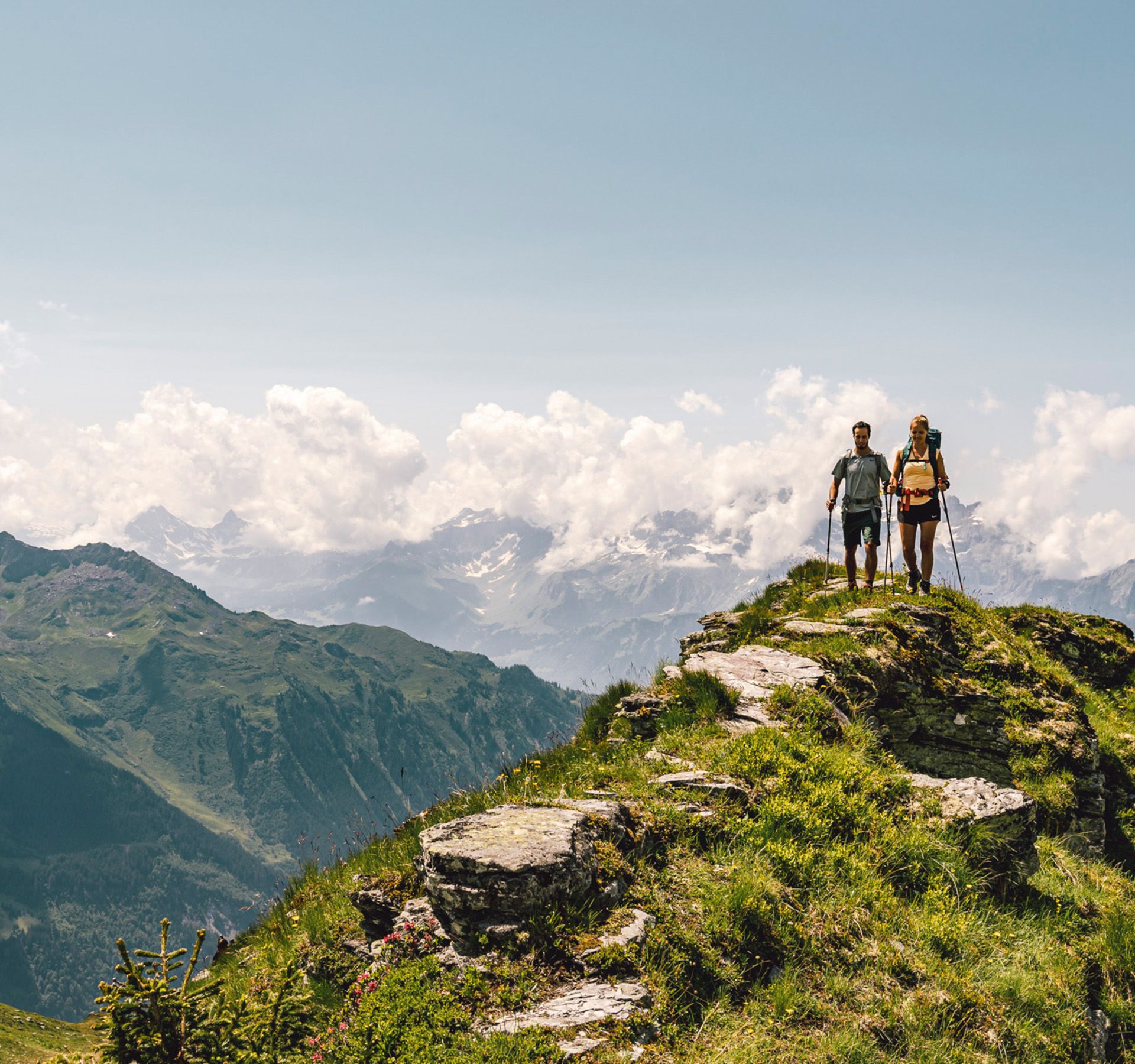 Mann und Frau wandern auf einem Grat bei schöne, sonnigen Wetter. Im Hintergrund sind zahlreiche, wolkenverhangene und teilweise schneebedeckte Berggipfel zu sehen.