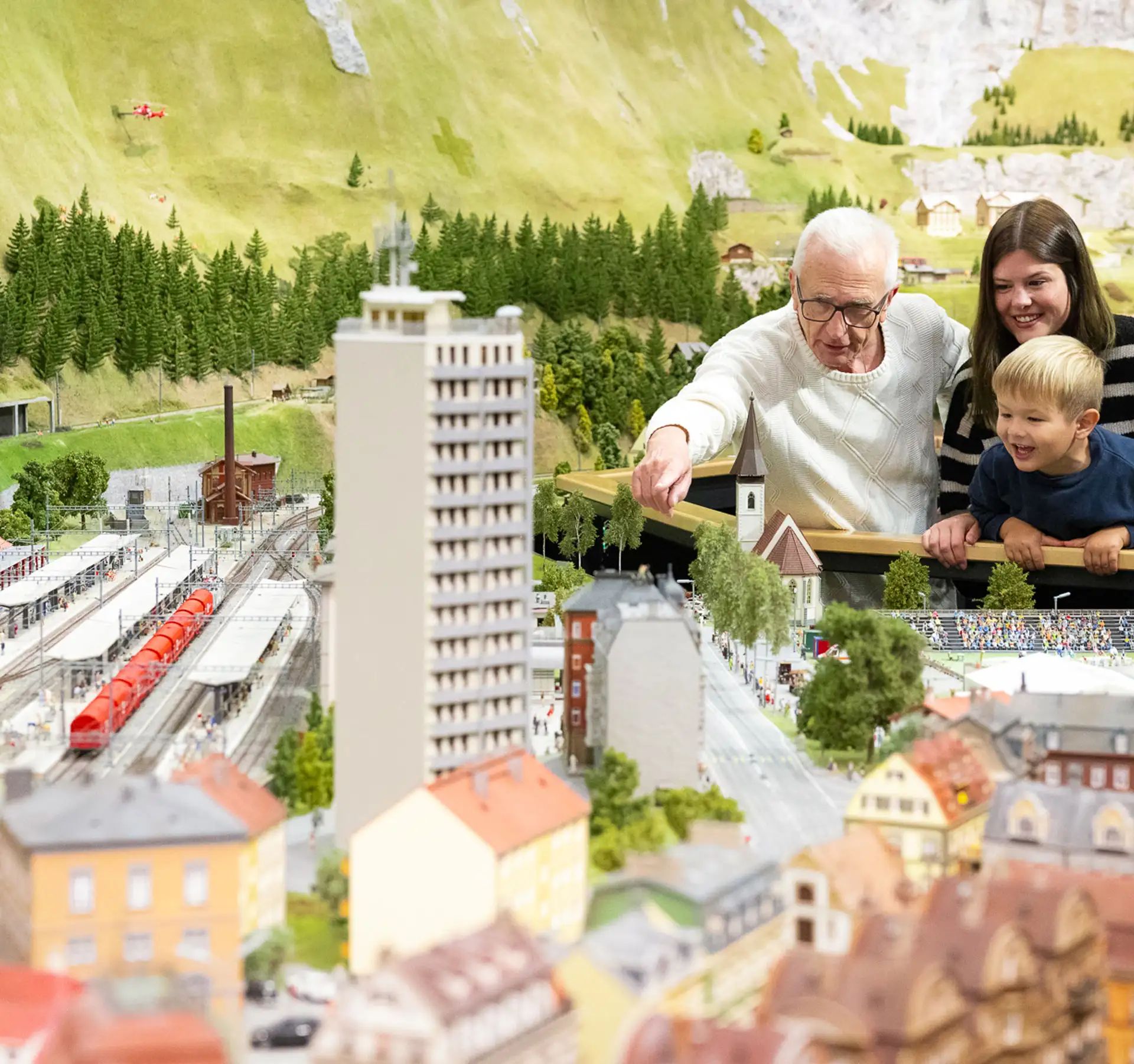 Several people observing a detailed miniature world with buildings, trains, and mountains in the background