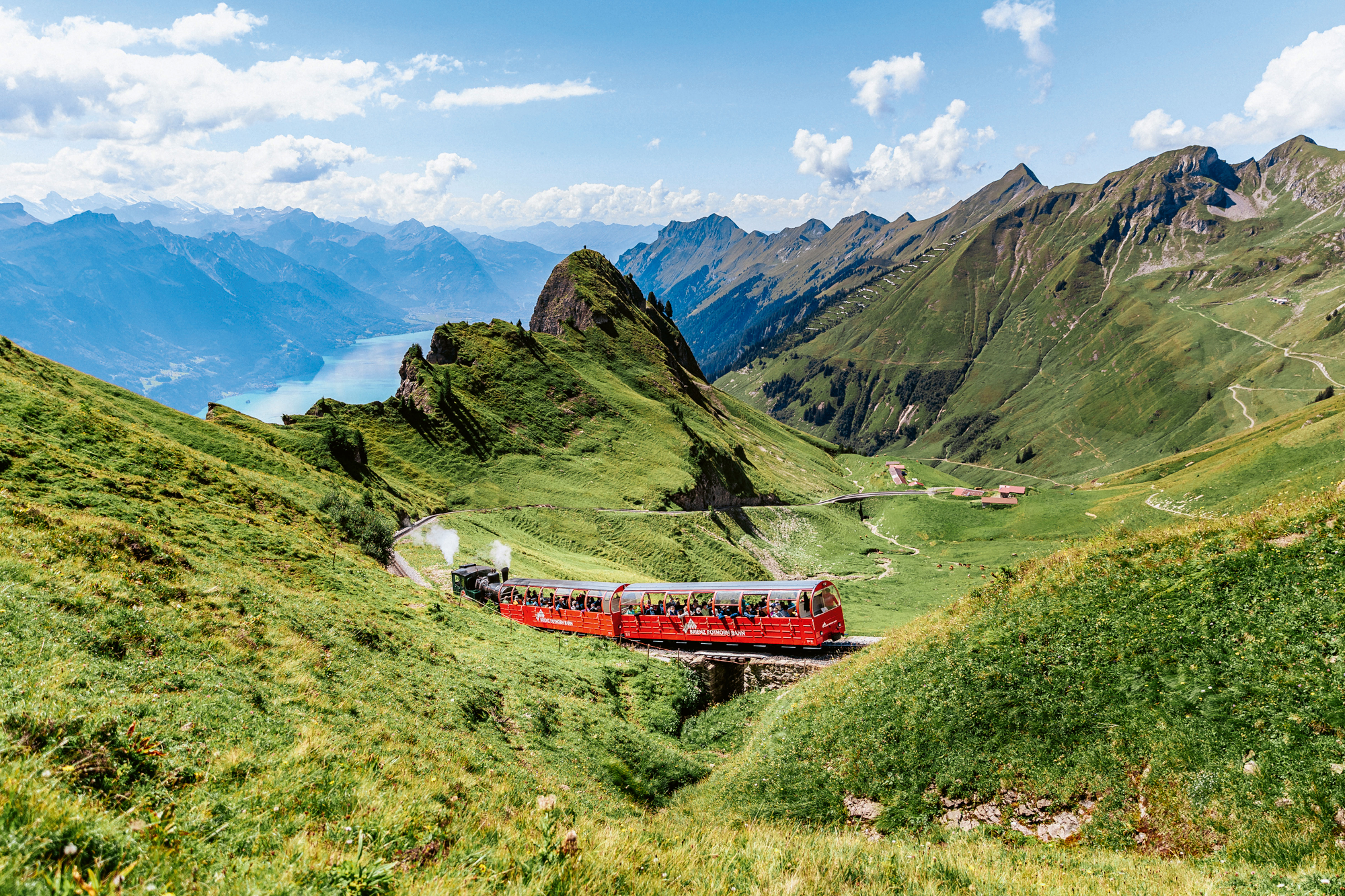 La locomotiva a vapore sale sul Brienzer Rothorn con le carrozze panoramiche rosse in una bella giornata estiva di sole. Il panorama offre una splendida vista sulla valle e sul blu intenso del lago di Brienz.