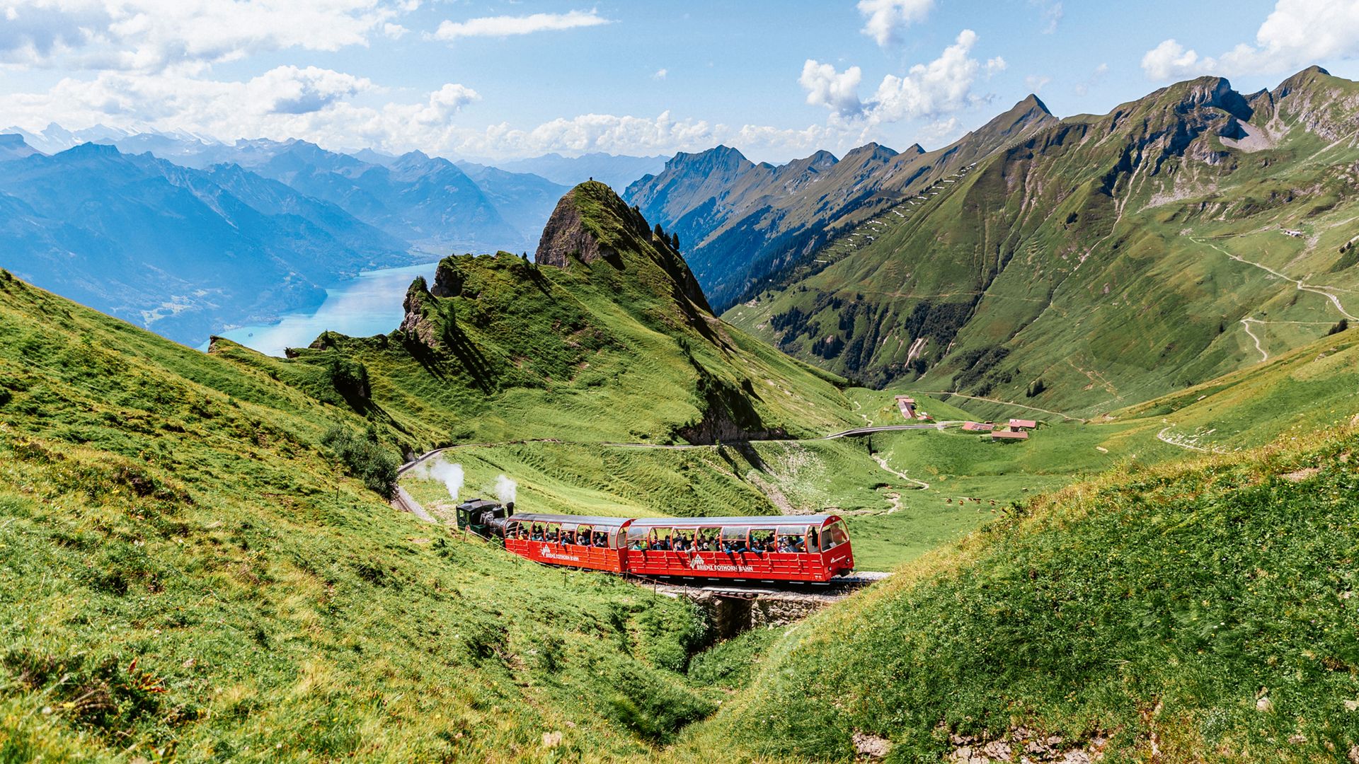 La locomotive à vapeur monte au Rothorn de Brienz avec des wagons panoramiques rouges par une magnifique journée d'été ensoleillée. Le panorama offre une vue magnifique jusqu'à la vallée et sur le lac de Brienz d'un bleu profond.