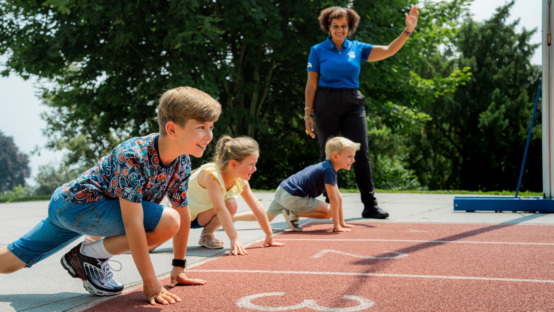 Three children at the starting line of a track, a woman signals the start.