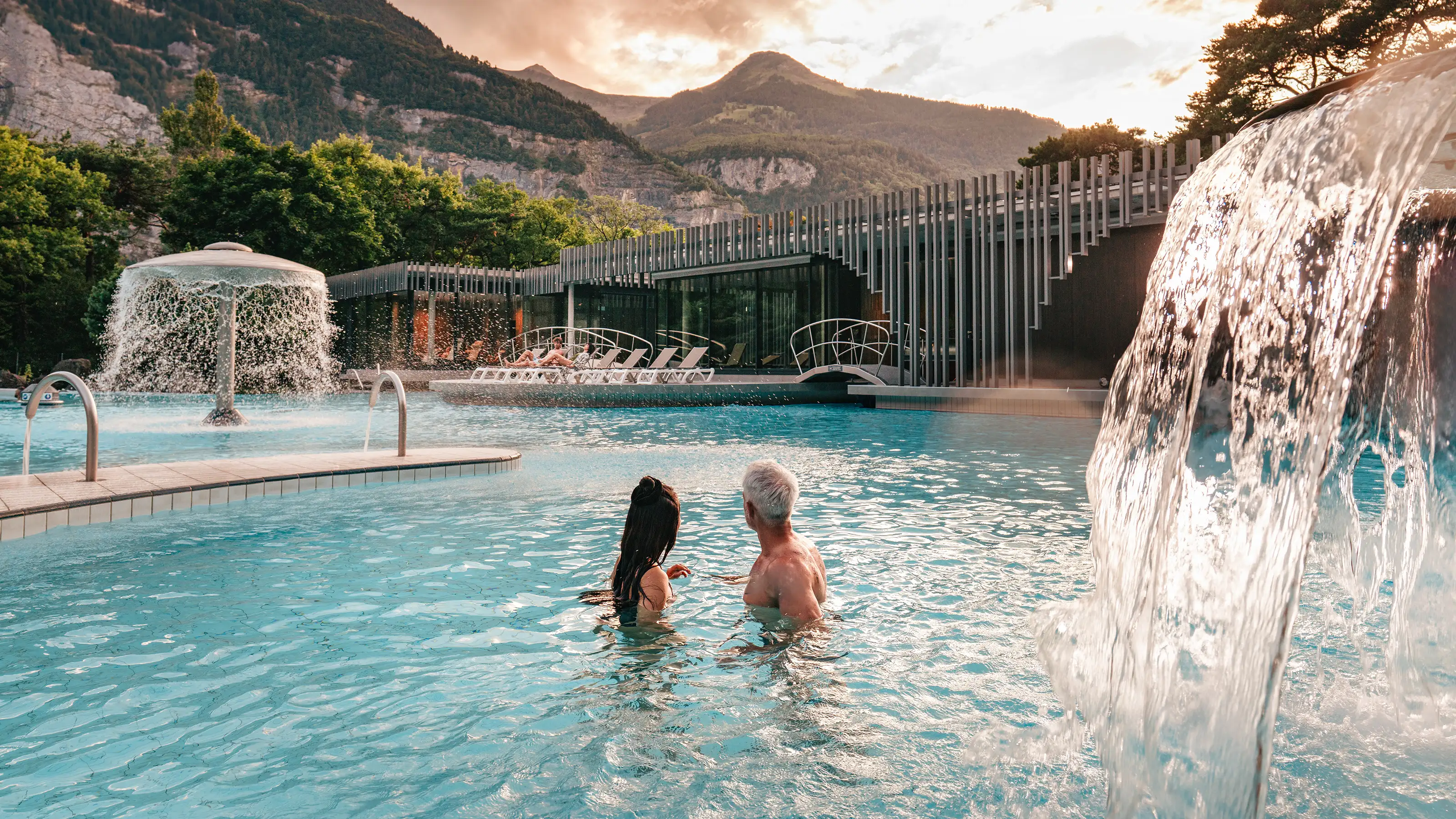 Deux personnes dans une piscine extérieure avec des cascades près d'un bâtiment moderne et des montagnes en arrière-plan.