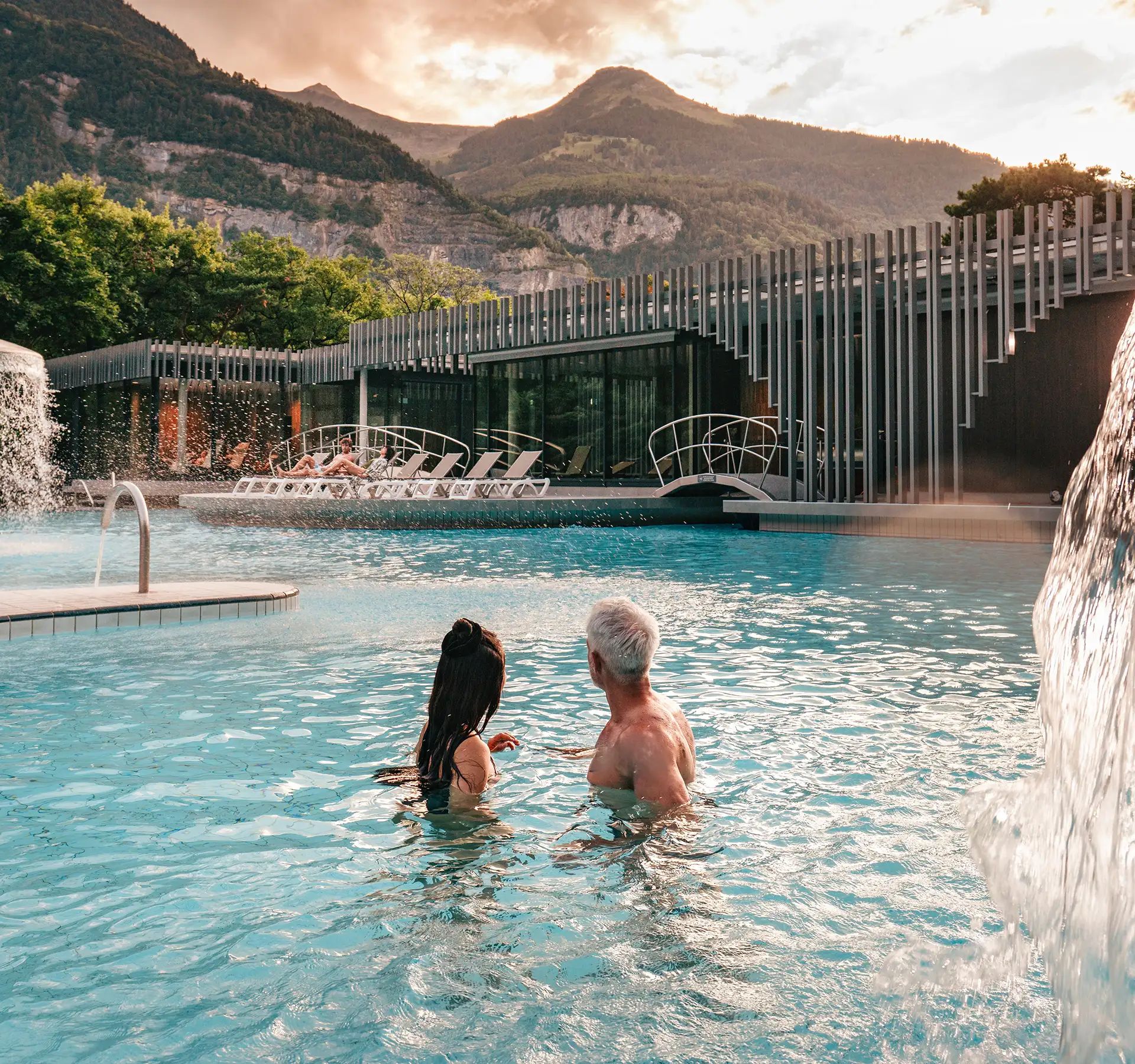 Deux personnes dans une piscine extérieure avec des cascades près d'un bâtiment moderne et des montagnes en arrière-plan.