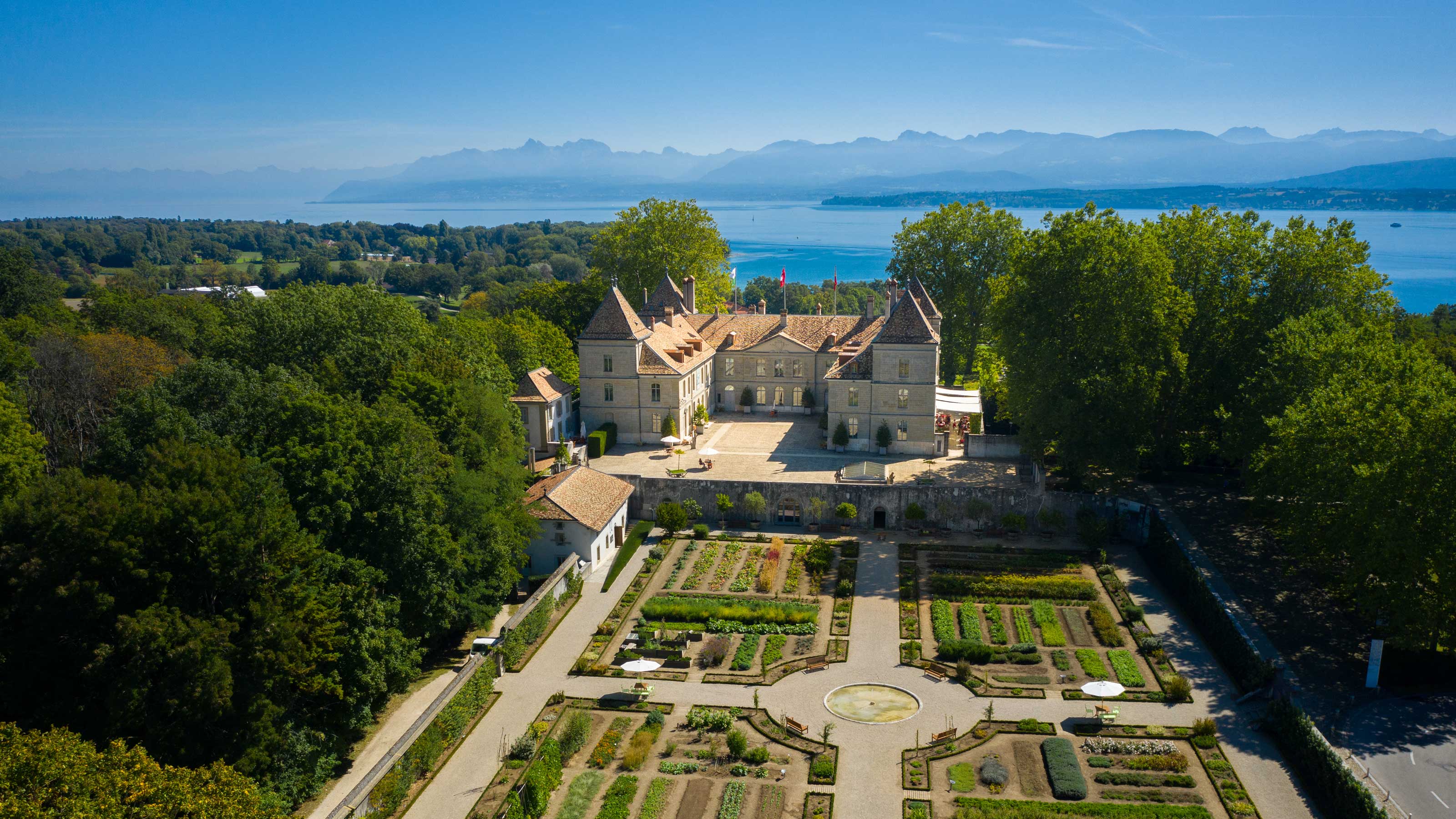Schloss mit einem grossen Garten auf der Vorderseite und Bergen und einem See im Hintergrund.