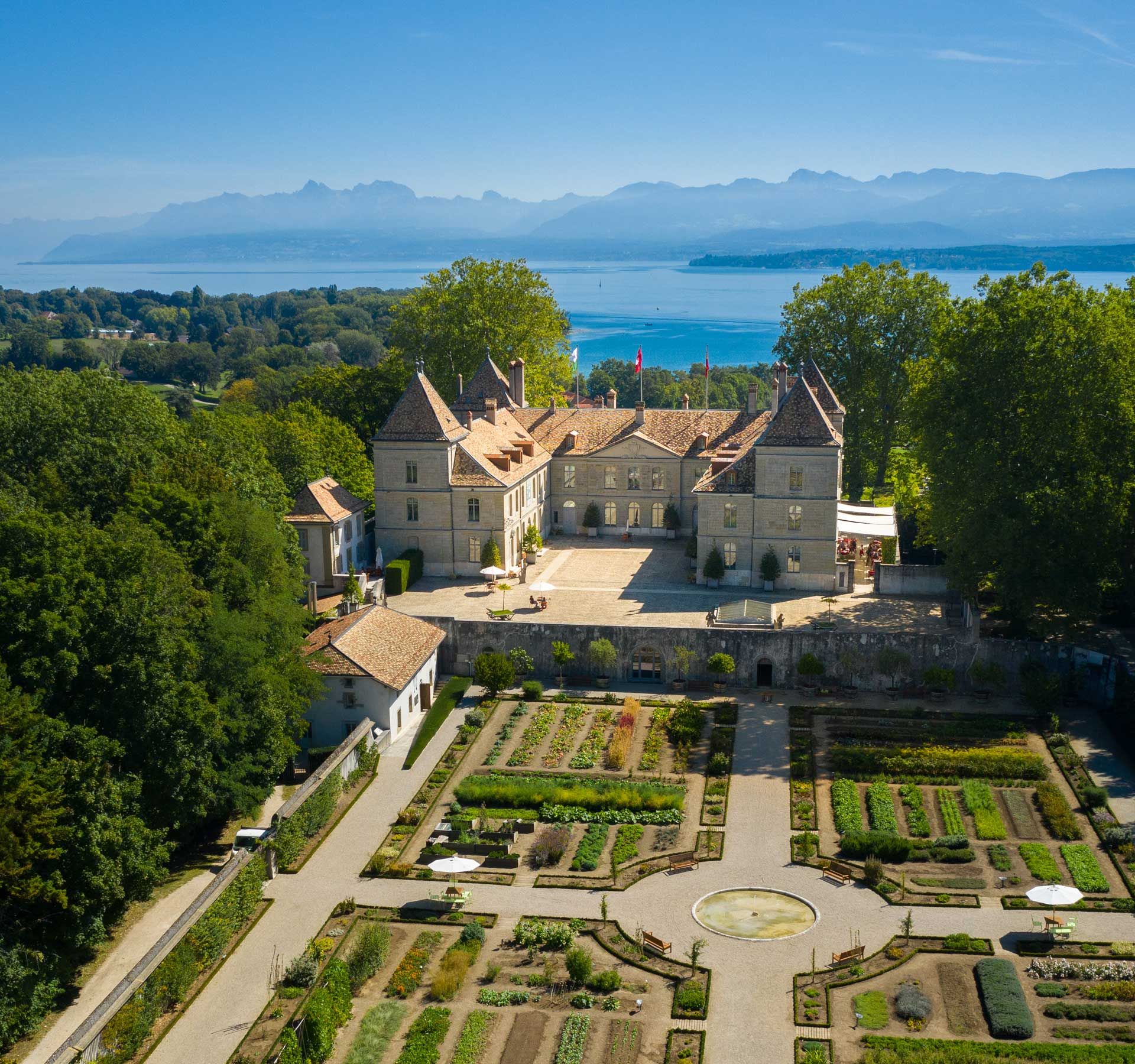 Castle with a large garden at the front and mountains and a lake in the background.