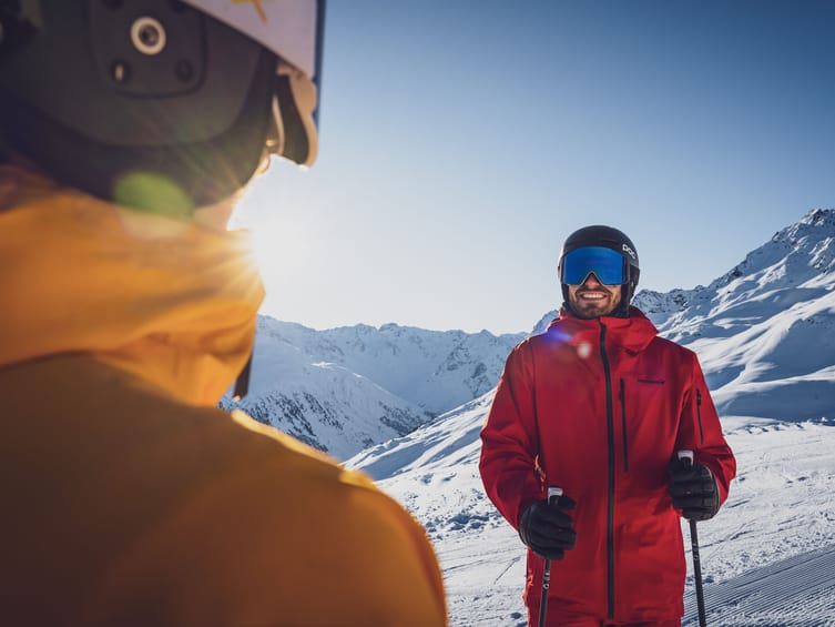 Un uomo con una giacca da sci rossa si trova su una montagna innevata. Sullo sfondo si vede un fantastico panorama montano con cime innevate.