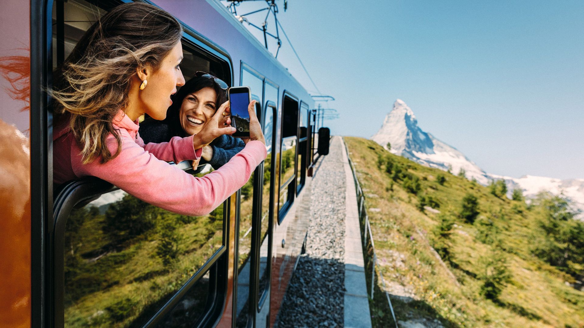 Two women lean slightly out of the window of the train and photograph the approaching Matterhorn above Zermatt. It is a glorious summer day with blue skies and sunshine. 