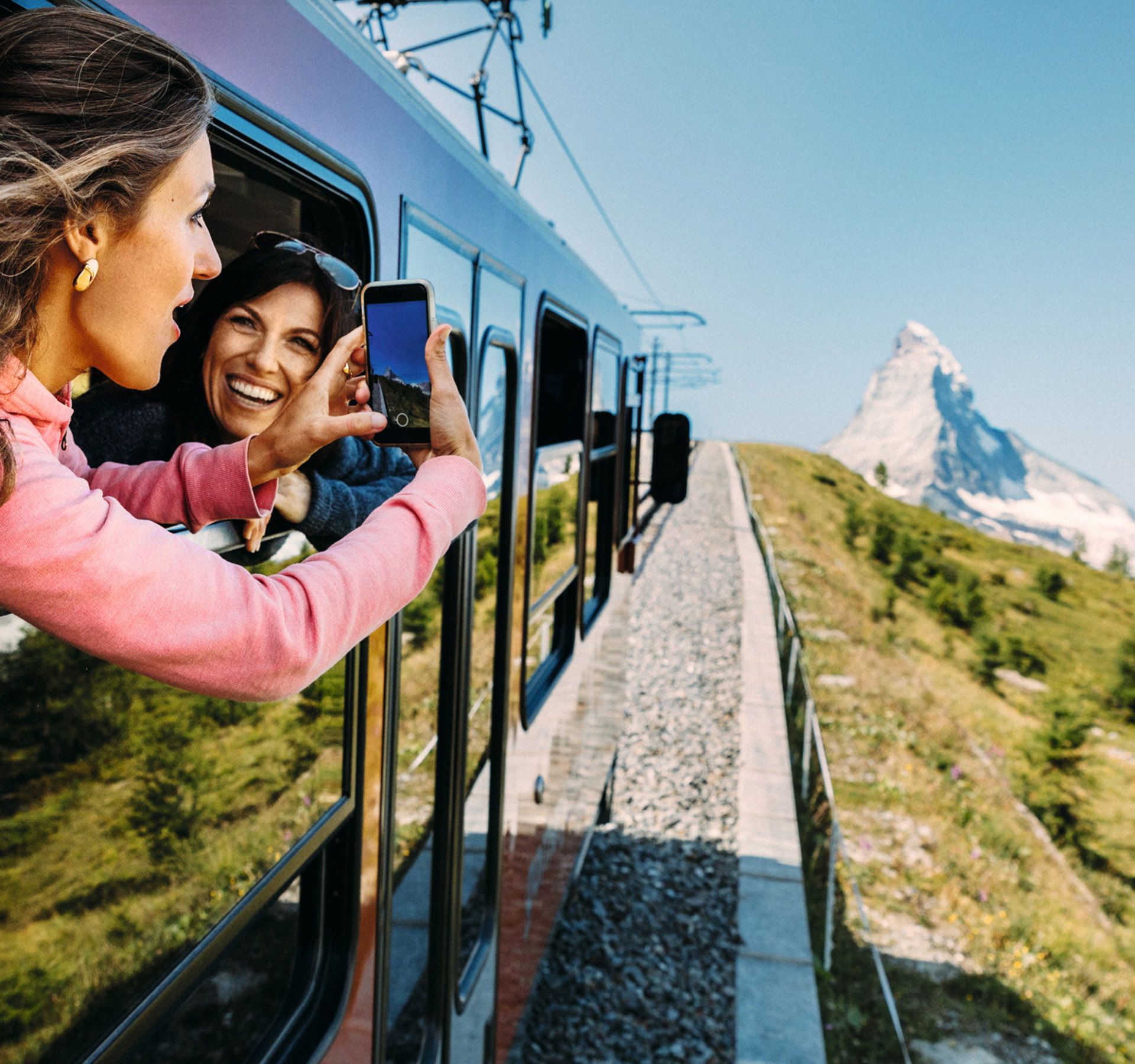 Two women lean slightly out of the window of the train and photograph the approaching Matterhorn above Zermatt. It is a glorious summer day with blue skies and sunshine.