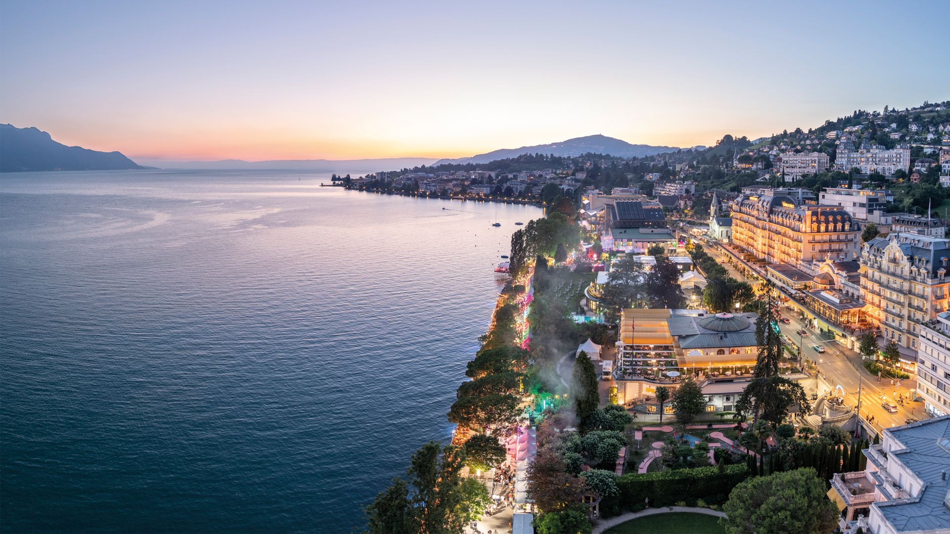 Blick auf die Uferpromenade von Montreux am Genfersee bei Sonnenuntergang mit beleuchteten Gebäuden und Bergen im Hintergrund.