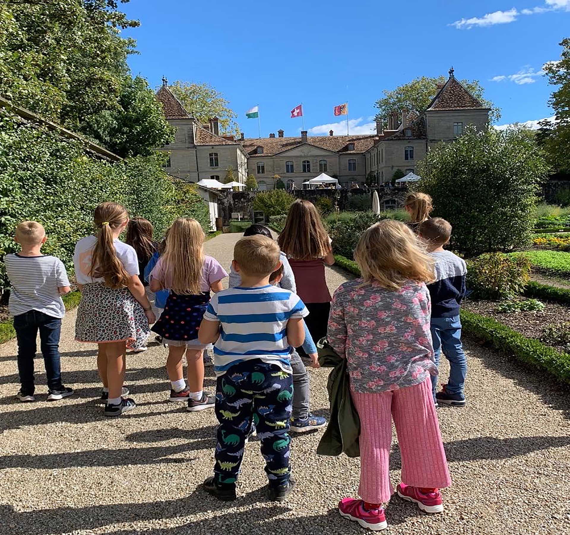 Un groupe d'enfants dans un jardin, regardant le château de Prangins avec des drapeaux en arrière-plan.