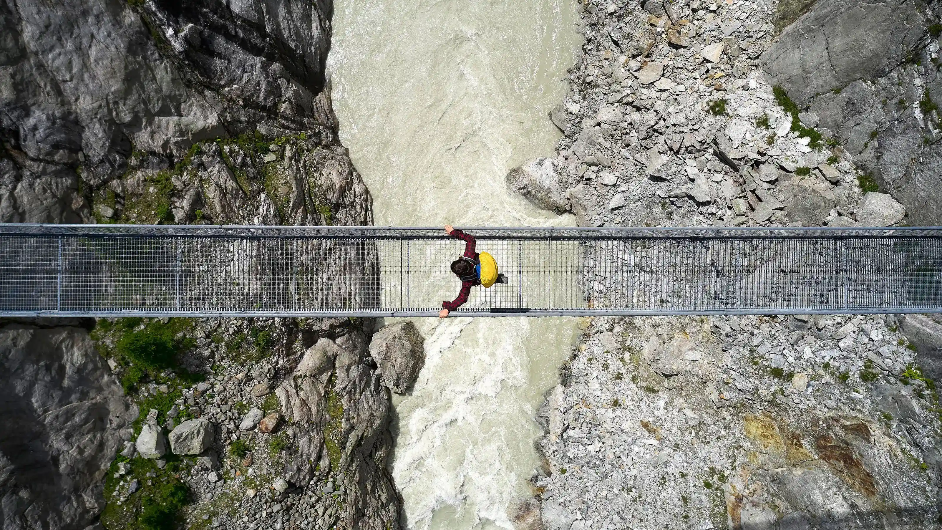  Persona con camicia rossa e zaino giallo attraversa ponte sospeso sopra fiume impetuoso in gola rocciosa
